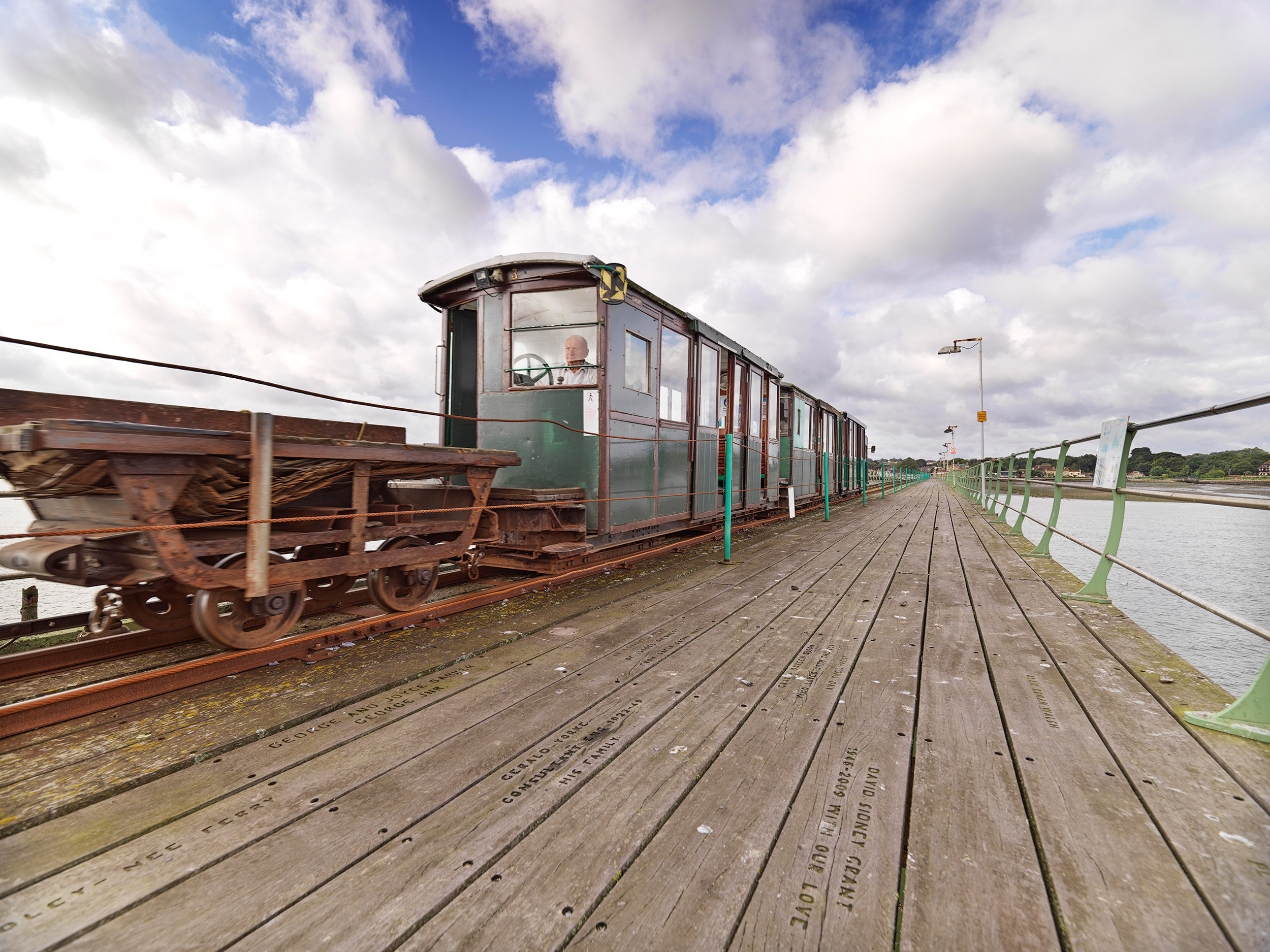 Photo along wooden pier with train on the tracks on the left and iron railing running down the right hand side.