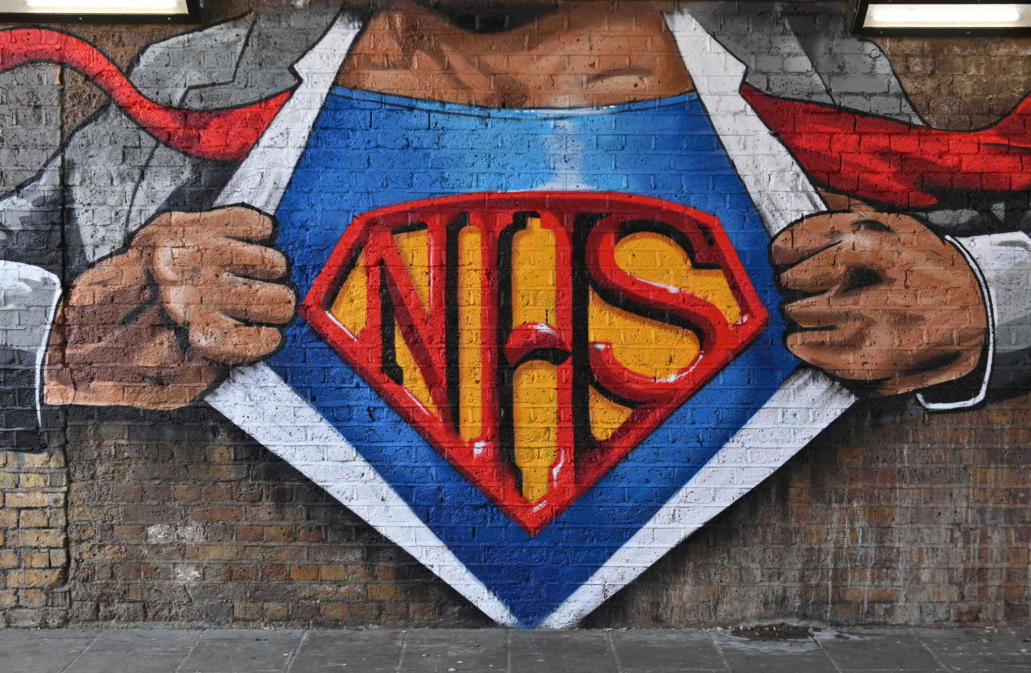 Work by a graffiti artist depicting the torso of a superman-style character pulling back his shirt to reveal a costume bearing the letters ‘NHS’.