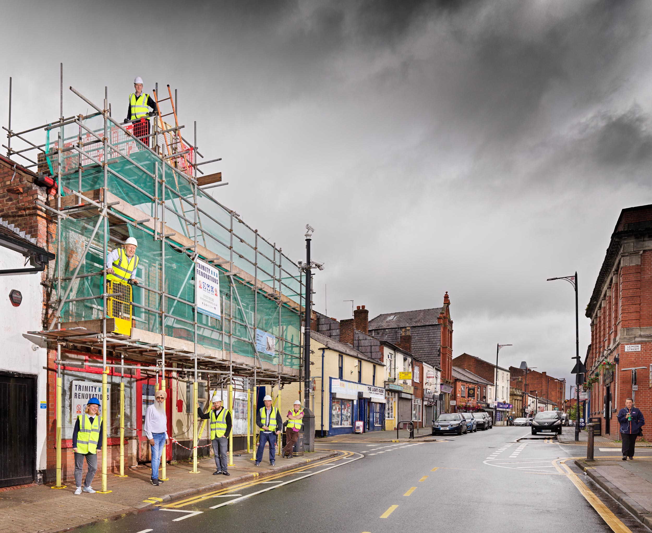 On the left side of the image six adults wearing hard hats and high visibility jackets and one man wearing a turban pose for a photo on and under three levels of scaffolding. The photo is taken from the middle of Elliott Street down its length.