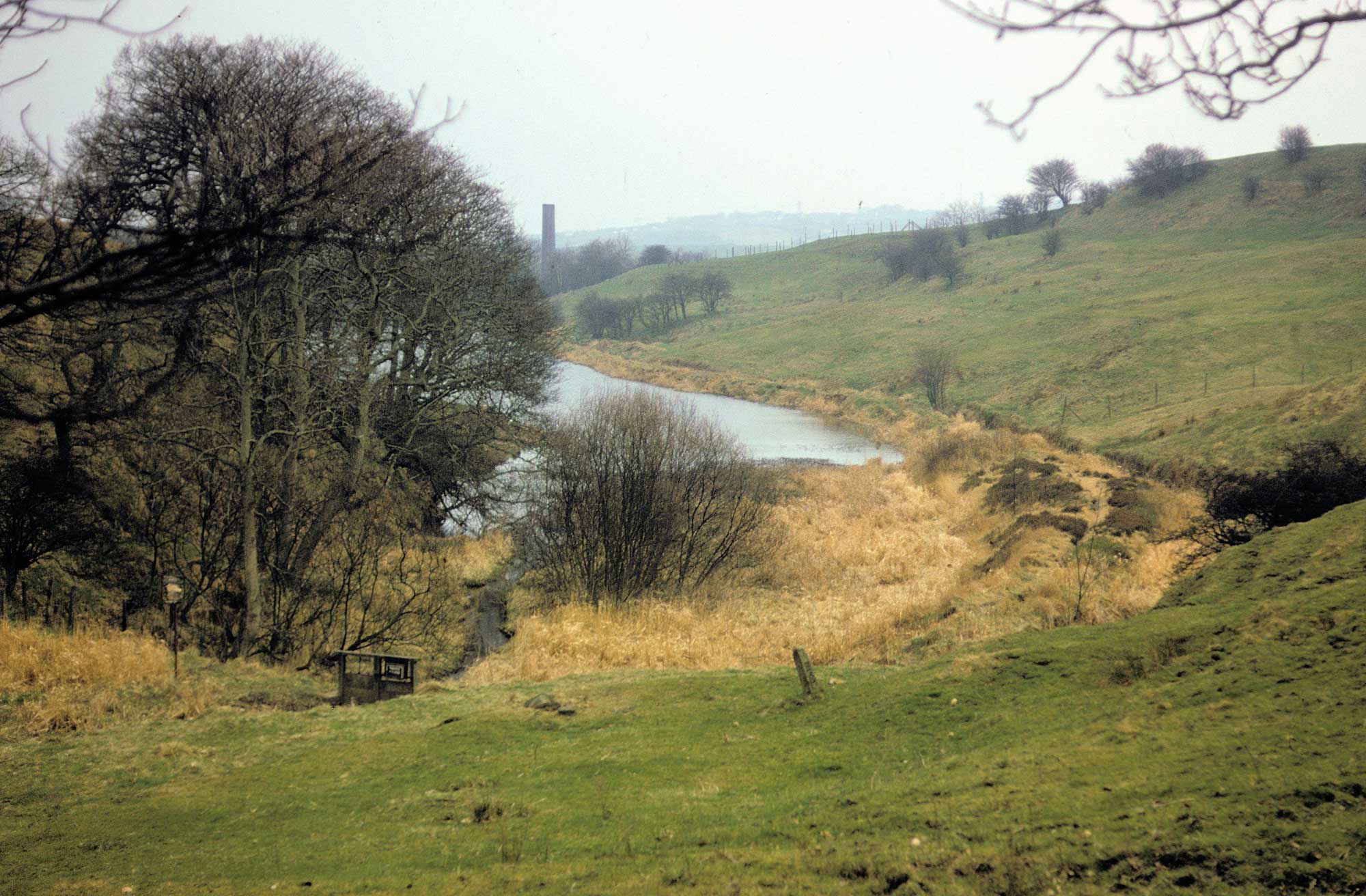 Reservoir situated between rolling hills. Industrial works are visible in the background