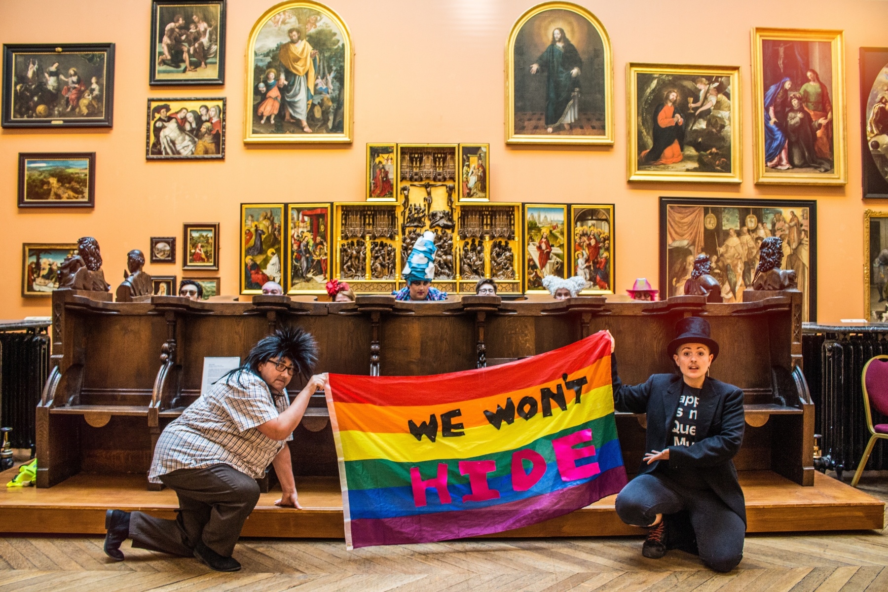 9 drag performers of various ages, genders and ethnicities pose in a large museum room with religious paintings on the walls. 7 of the performers are hiding behind an ornate wooden structure in the centre of the room, with only their heads showing. 2 of the performers keel in front of the structure holding a rainbow flag into which the following text has been sewn in black and pink lettering "We Won't Hide"