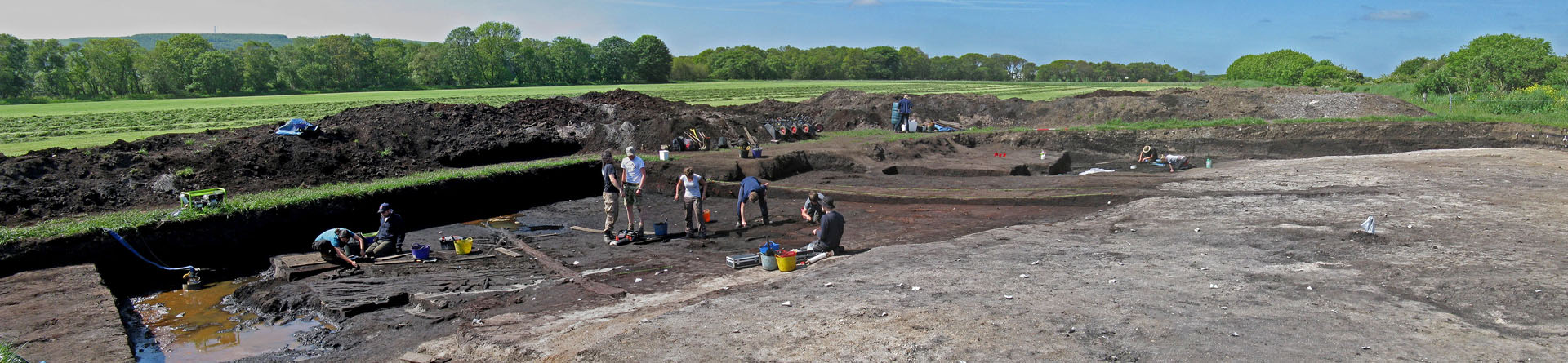 Colour photograph showing lots of archaeologists digging in a trench with spoilheaps behind; some timbers are visible left