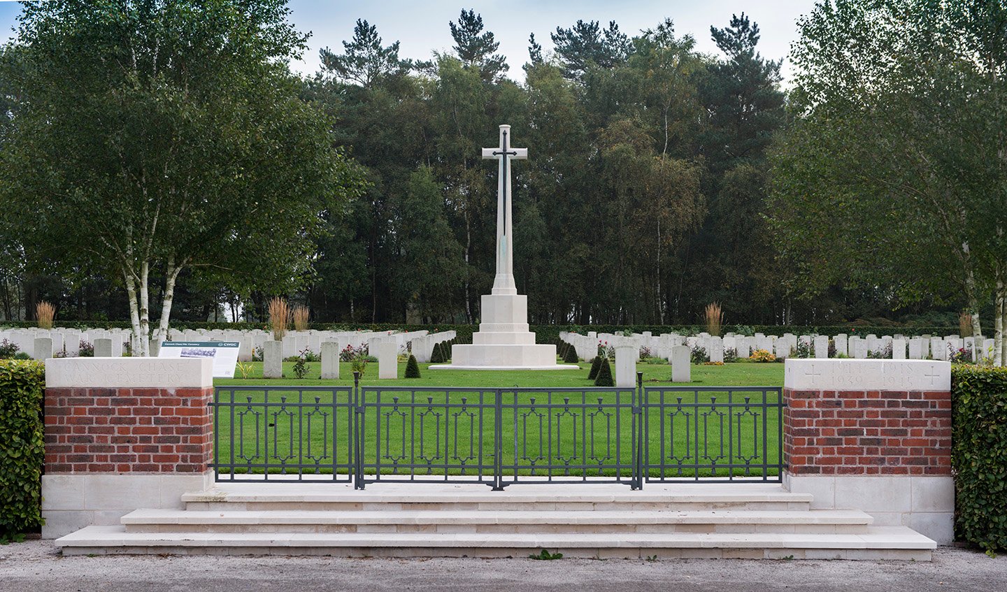 Colour photograph showing a cenotaph behind a metal gate with rows of well tended gravestones in the background