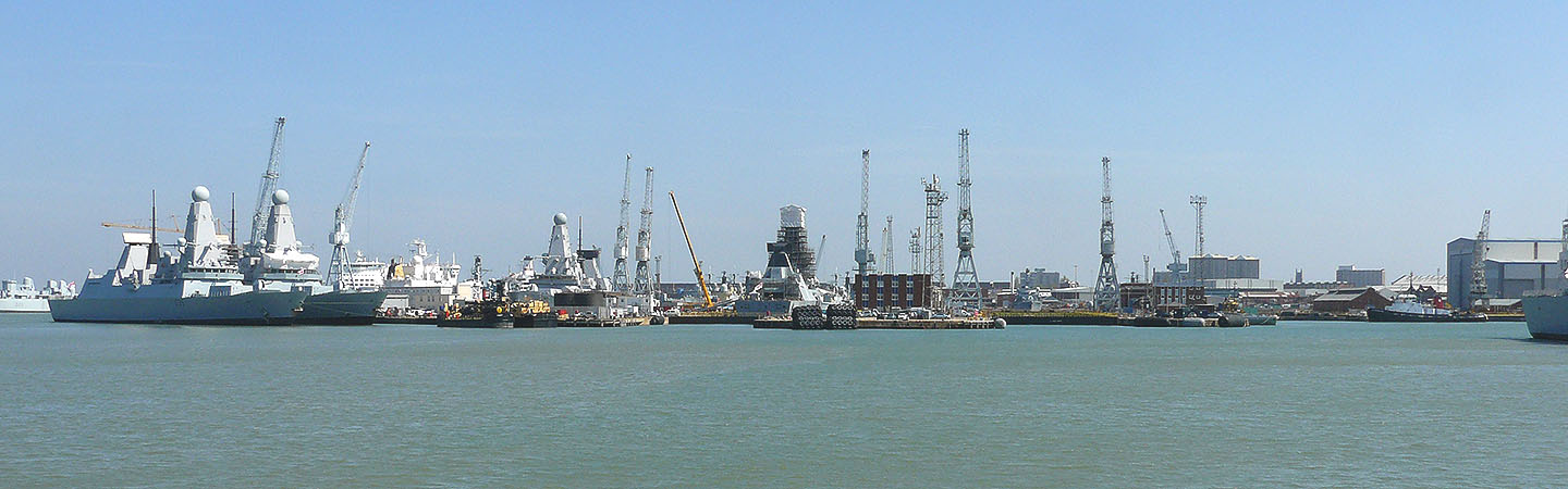 Alt txt – Portsmouth Dockyard, Hampshire, moored warships in the dockyard with cranes to the rear.