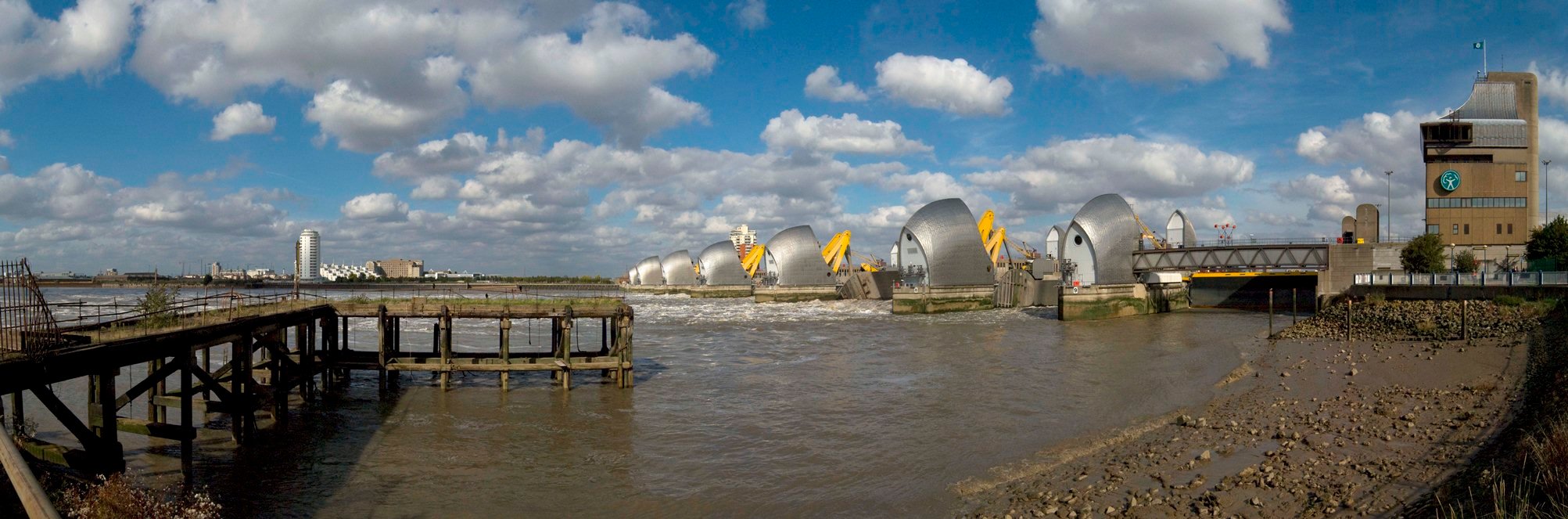 A wide angled photograph of a jetty and rocky foreshore in front of the Thames Barrier, London.