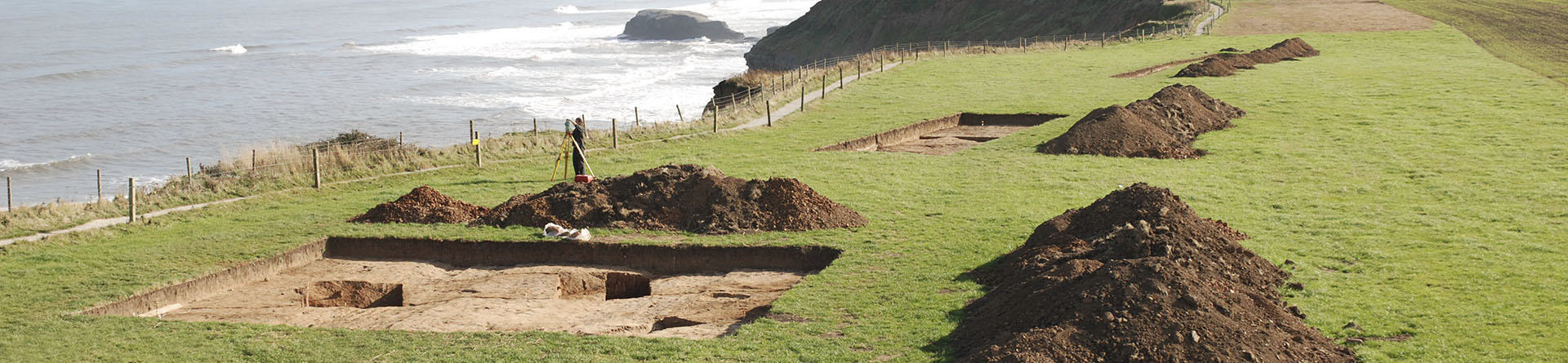 Piles of soil next to excavated trenches on the cliffs edge with the sea in the background