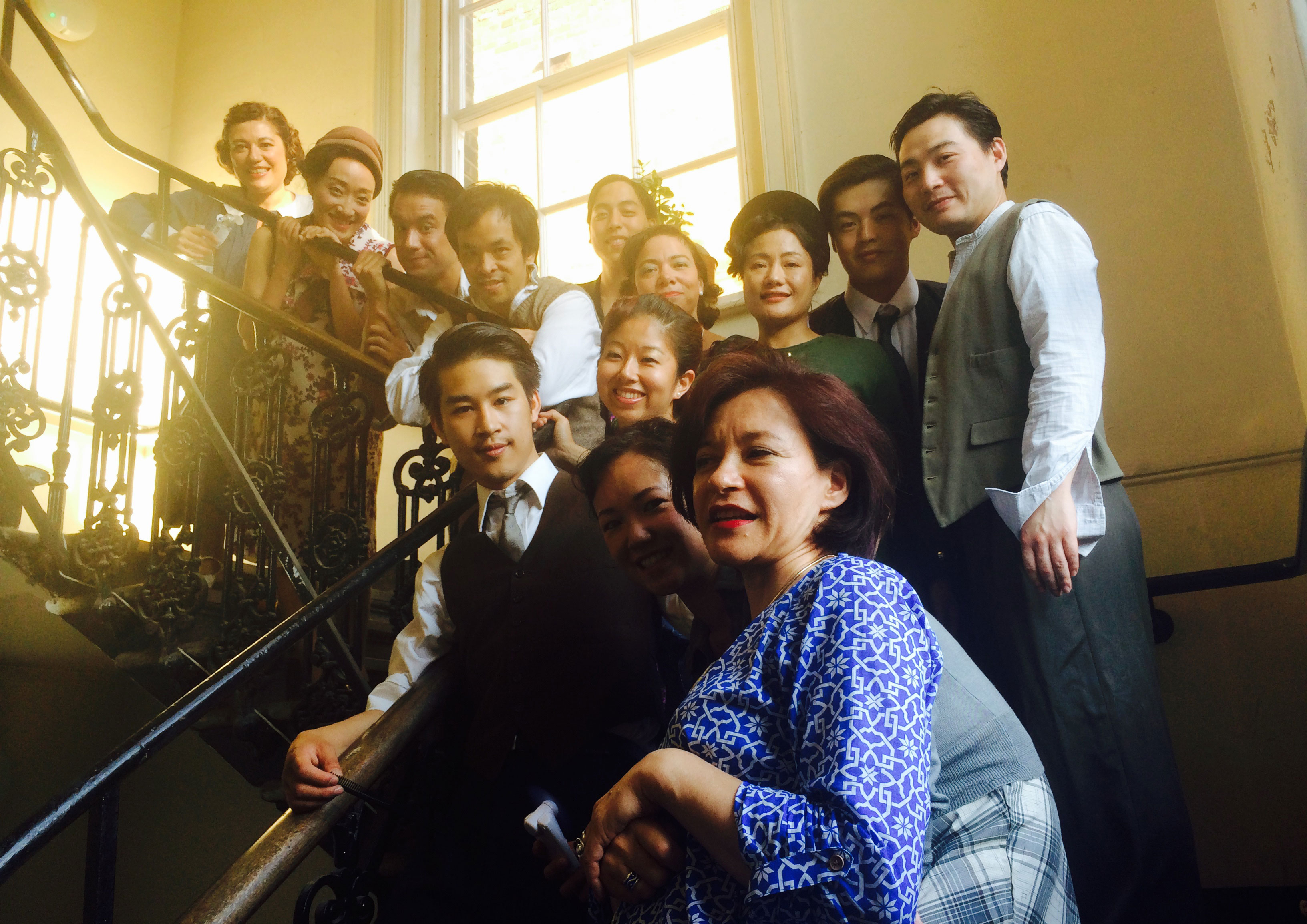 Kumiko Mendl with the cast of ‘The Last Days of Limehouse’ posing for a group photo on a stairway with wrought iron banisters.