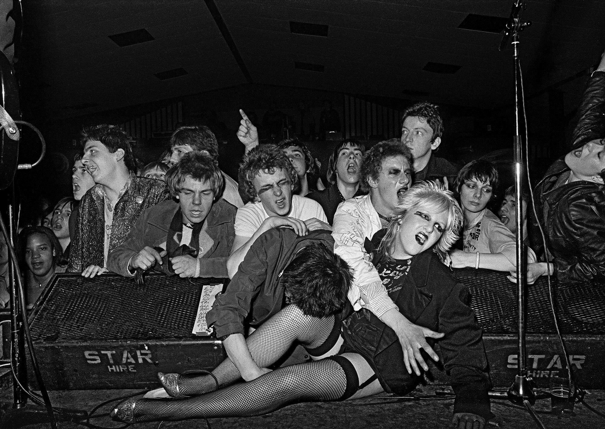 Punk girl in fishnets and suspenders reclining between two monitors up on stage with other members of the gig audience behind and around her. Photo taken 1976.