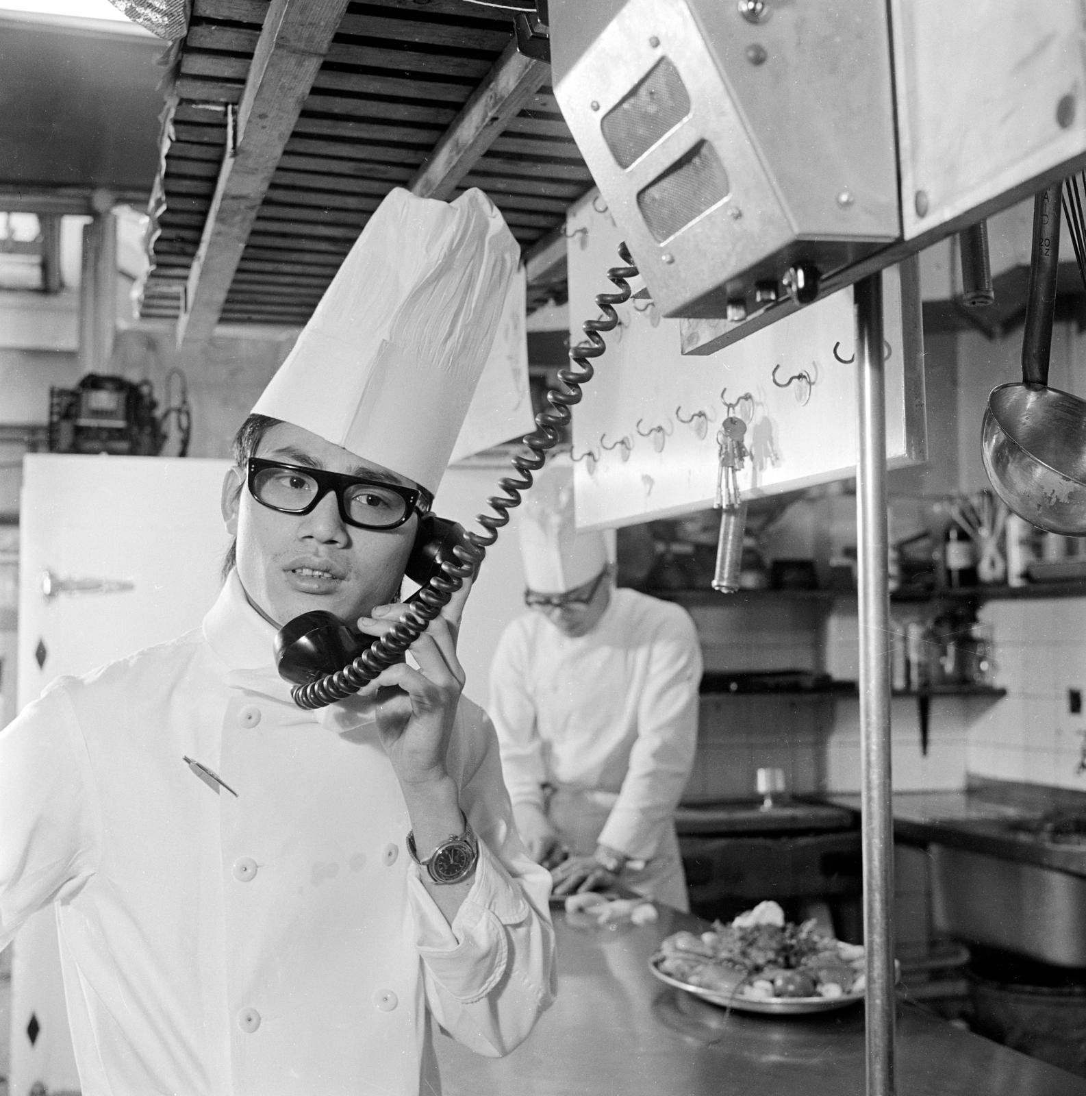 Chefs wearing whites at work in the kitchen at Wheelers Sovereign Restaurant. The chef in the foreground is talking on a telephone. Black and white photo taken 1960-1980.