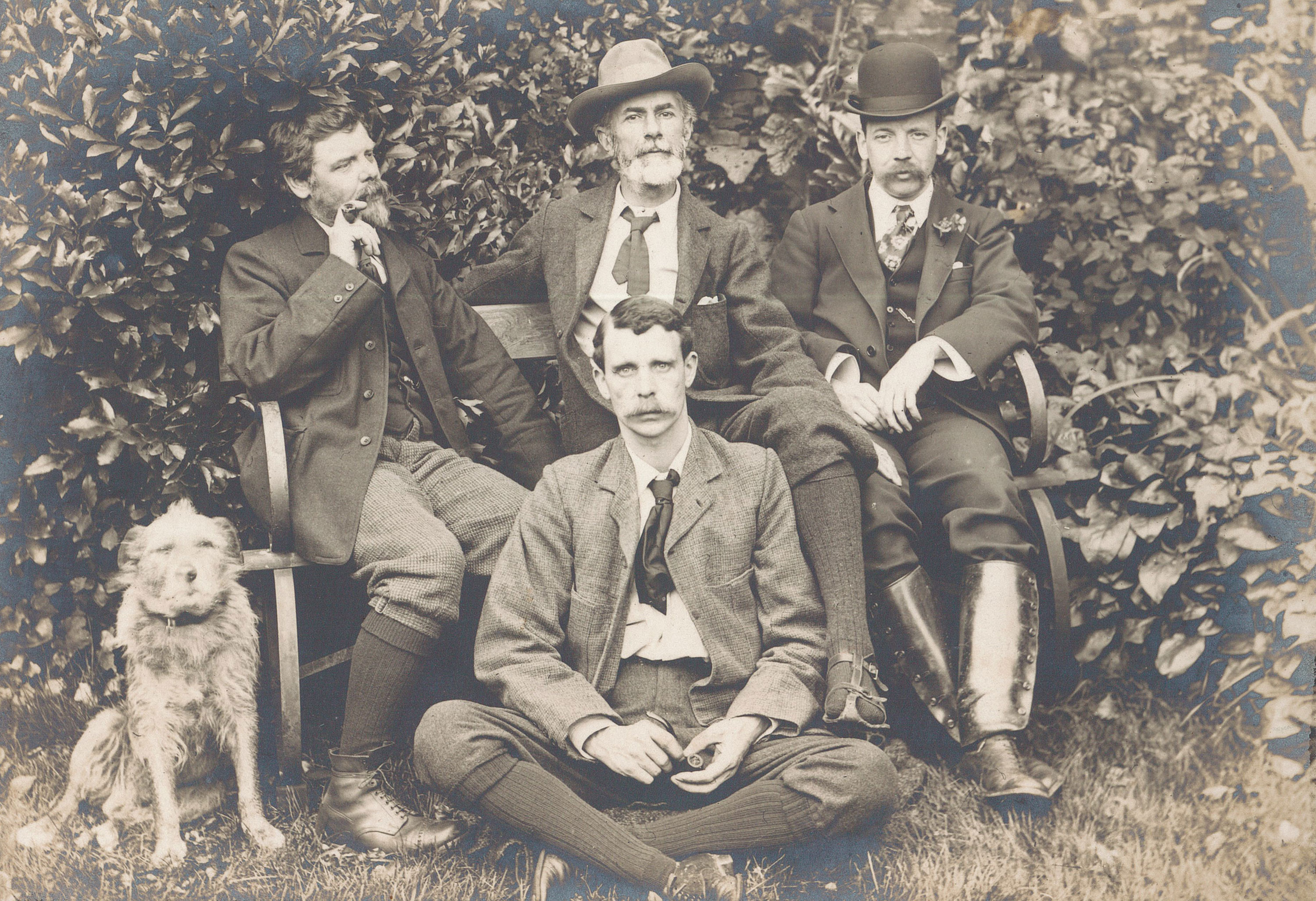 Sepia photo of Edward Carpenter, George Merril and Charles Sixsmith seated on a bench with G. Hukin and a dog sitting on the ground. Behind the bench is a high hedge.