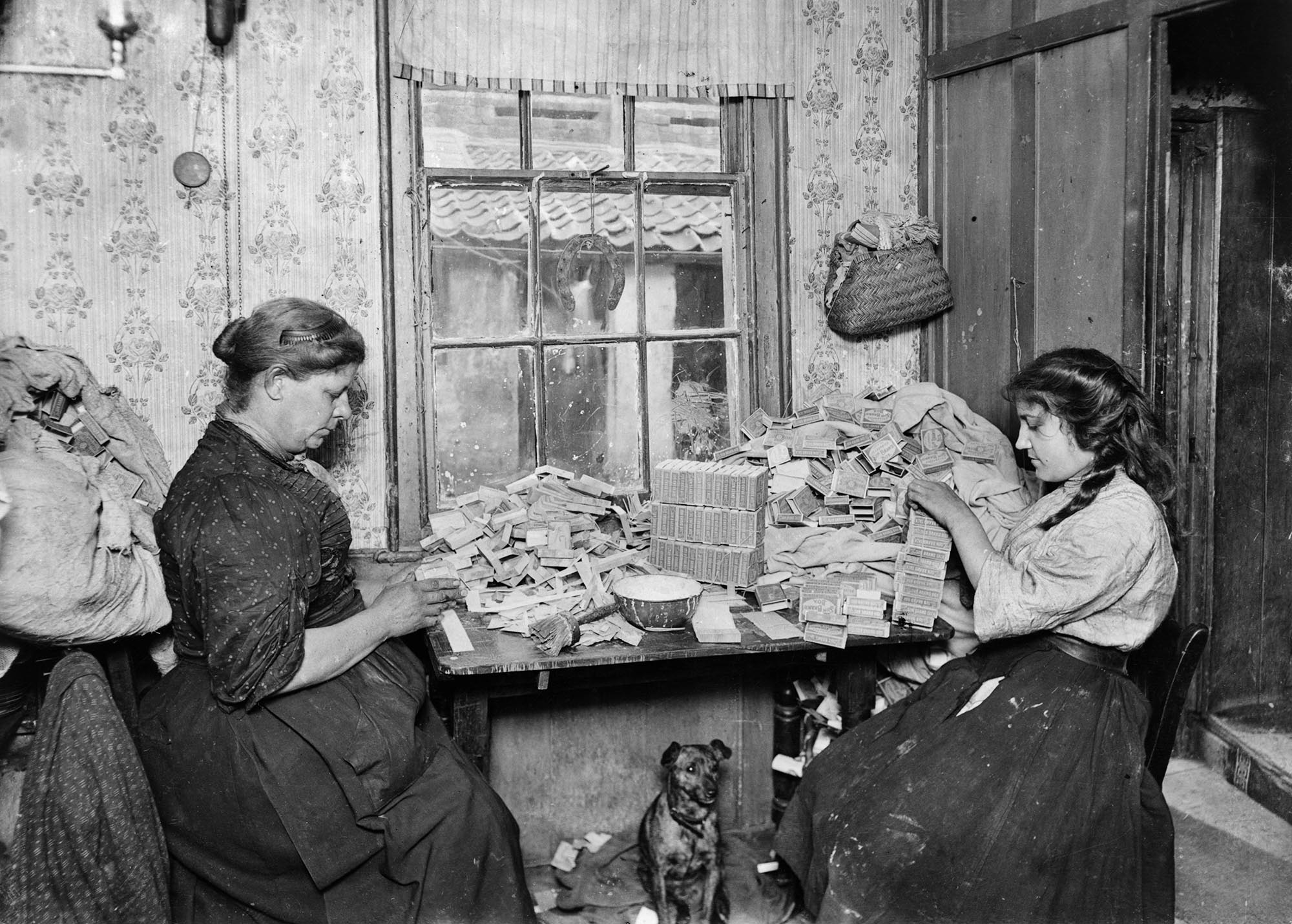 Two women assembling small boxes at a table in an unidentified house in the East End of London