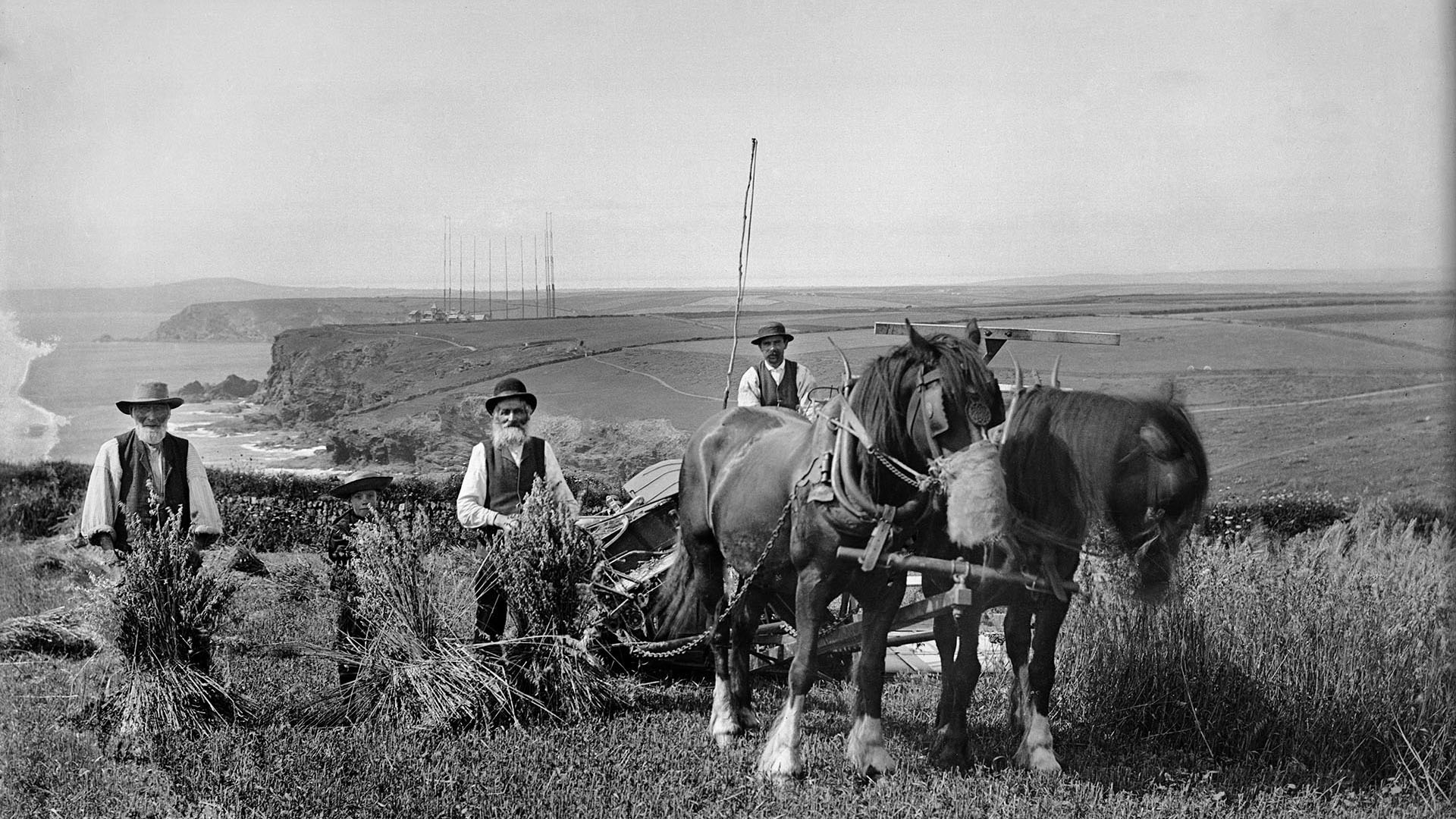 Black and white archive photograph of three farm workers and a horse drawn vehicle posing for the camera in a field, with radio masts, cliffs and coasts in the background.