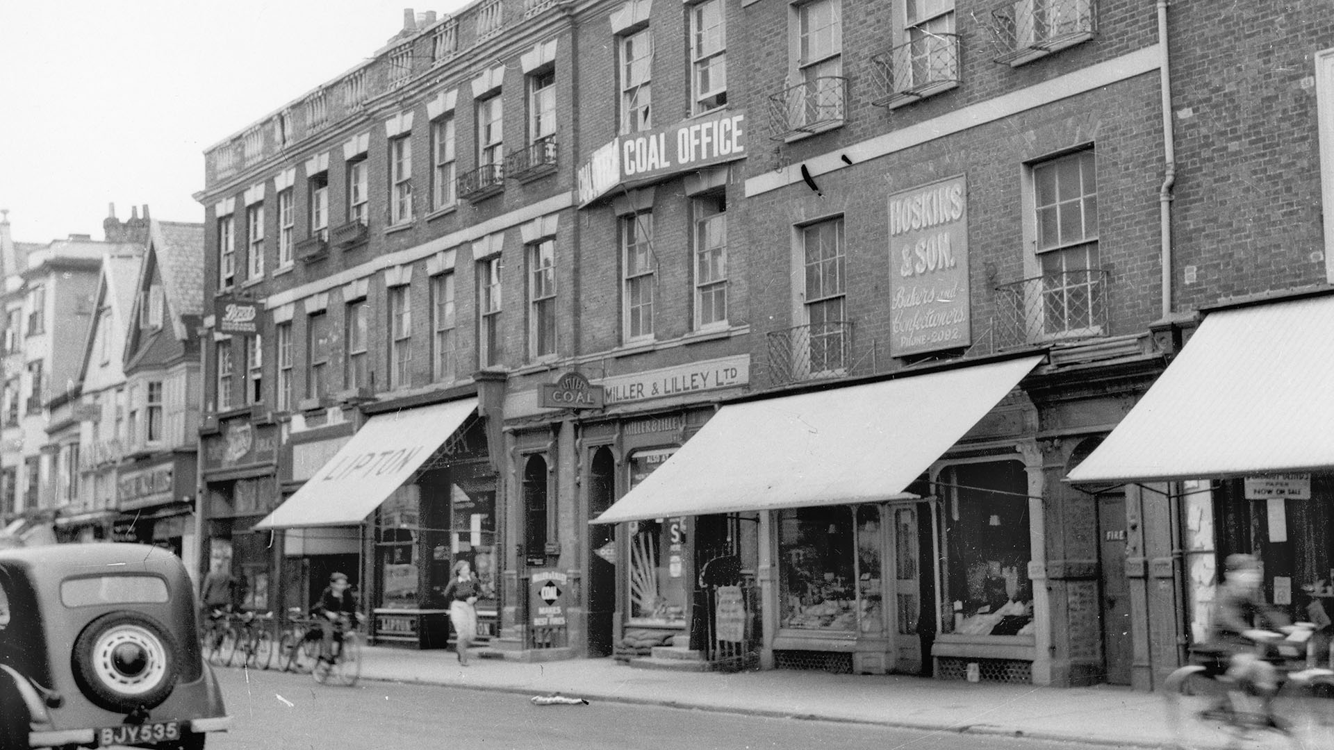 Black and white archive photograph of a row of shops in 1942, with a car, bicycles and a pedestrian.