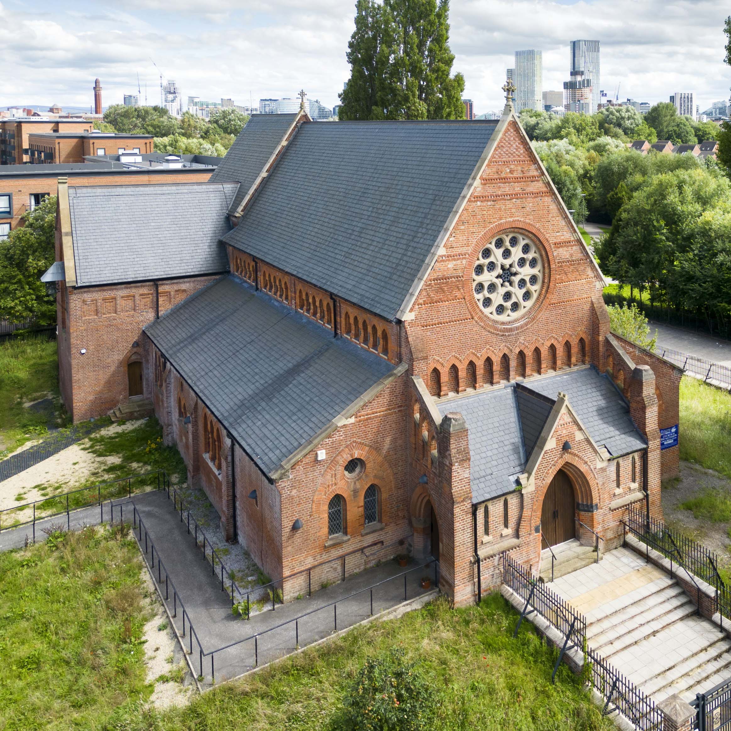 An exterior view of the west elevation of a Church from above with the Manchester skyline in the background. 