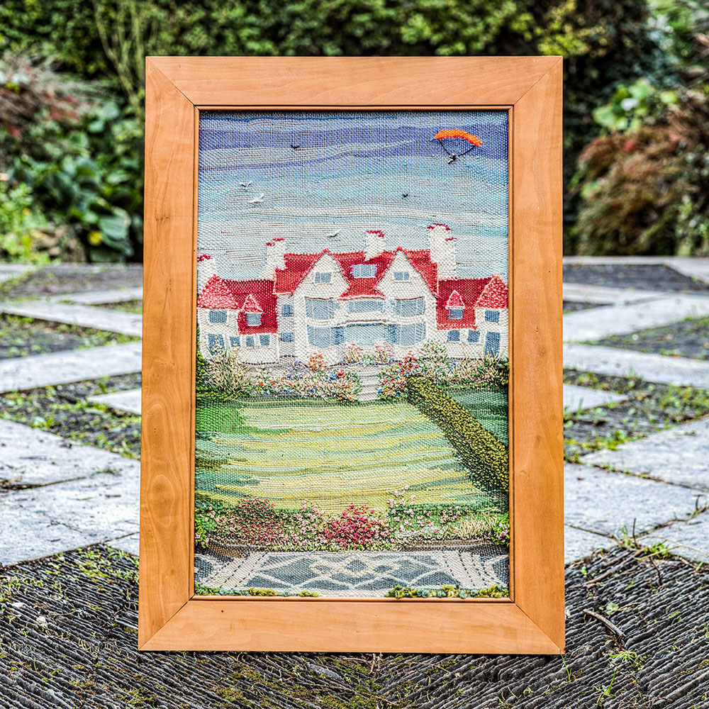 Image of an embroidered picture of the garden and lodge, sitting on the paving of the garden. 