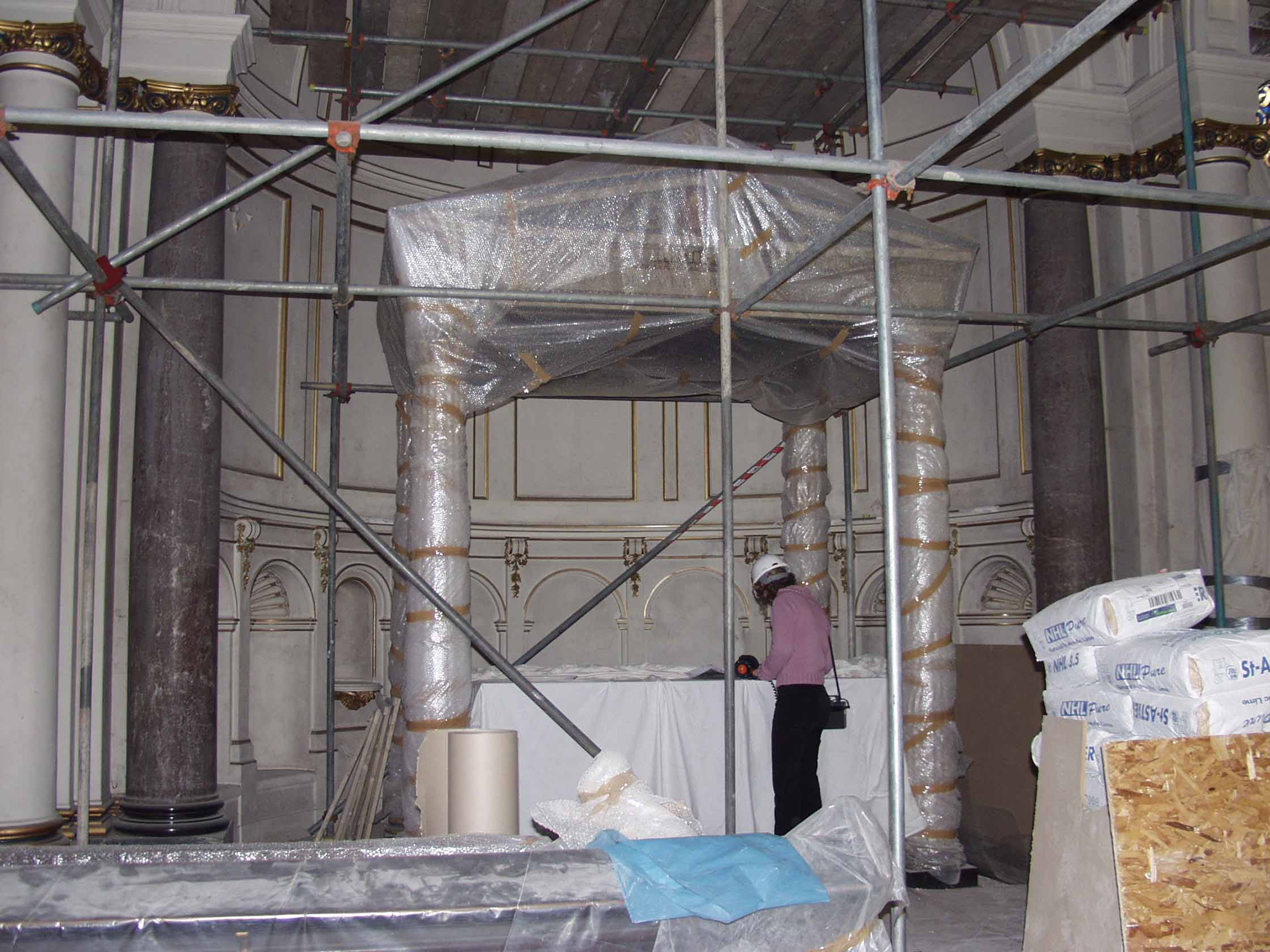 A person in a hard hat stands in front of a baldaquin covered in bubble-wrap, flanked by two columns and surrounded by scaffolding.