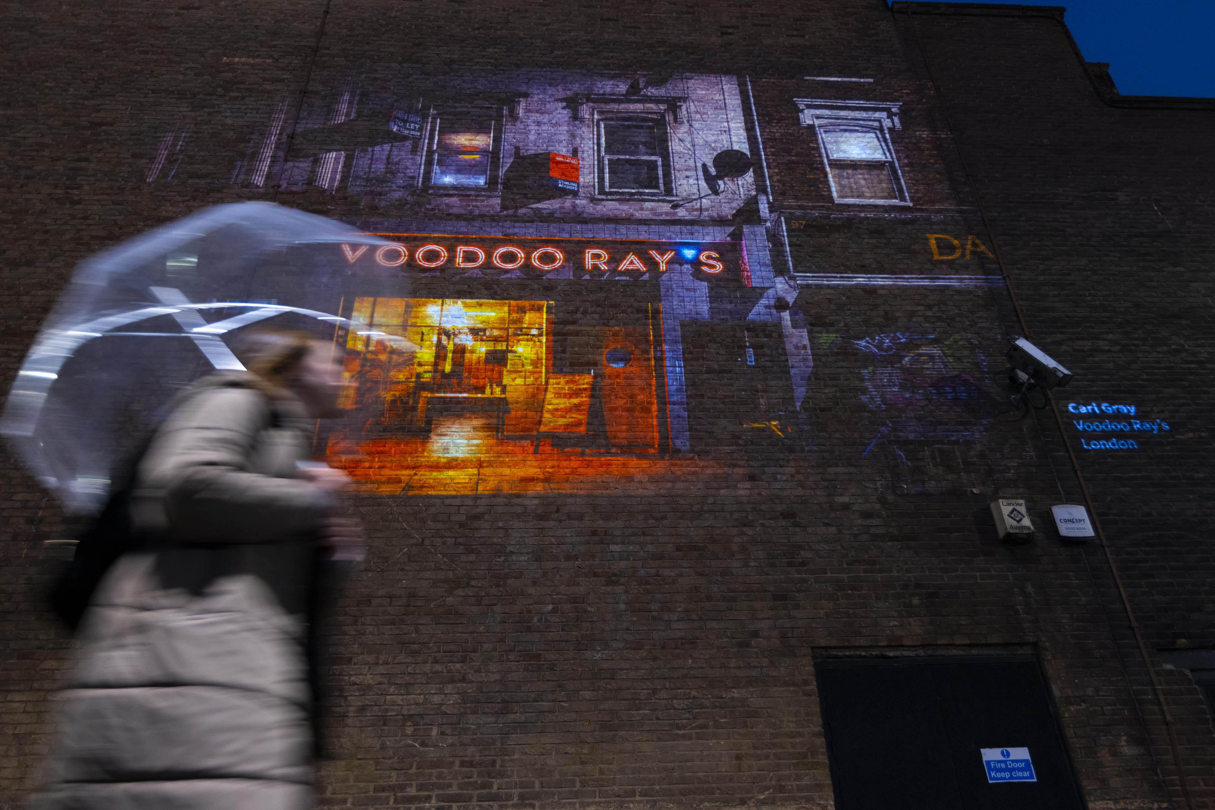 A photo by Carl Gray from Historic England’s ‘Picturing High Streets’ outdoor exhibition is projected outside The Photographers’ Gallery in London. A woman holding an umbrella is walking by.