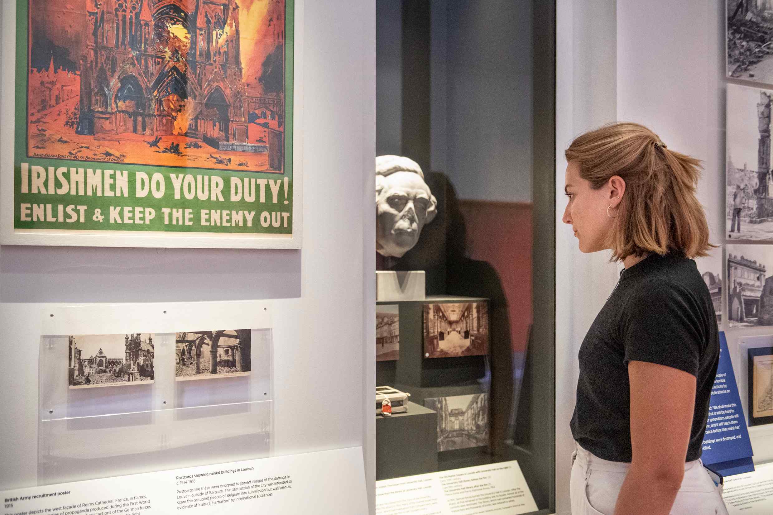 A woman visiting the What Remains exhibition looks into a display cabinet including a sculpture of a man's head that has had the nose knocked off. A poster to her left shows a church in flames and includes the text: "IRISHMEN DO YOUR DUTY! Enlist and keep the enemy out"
