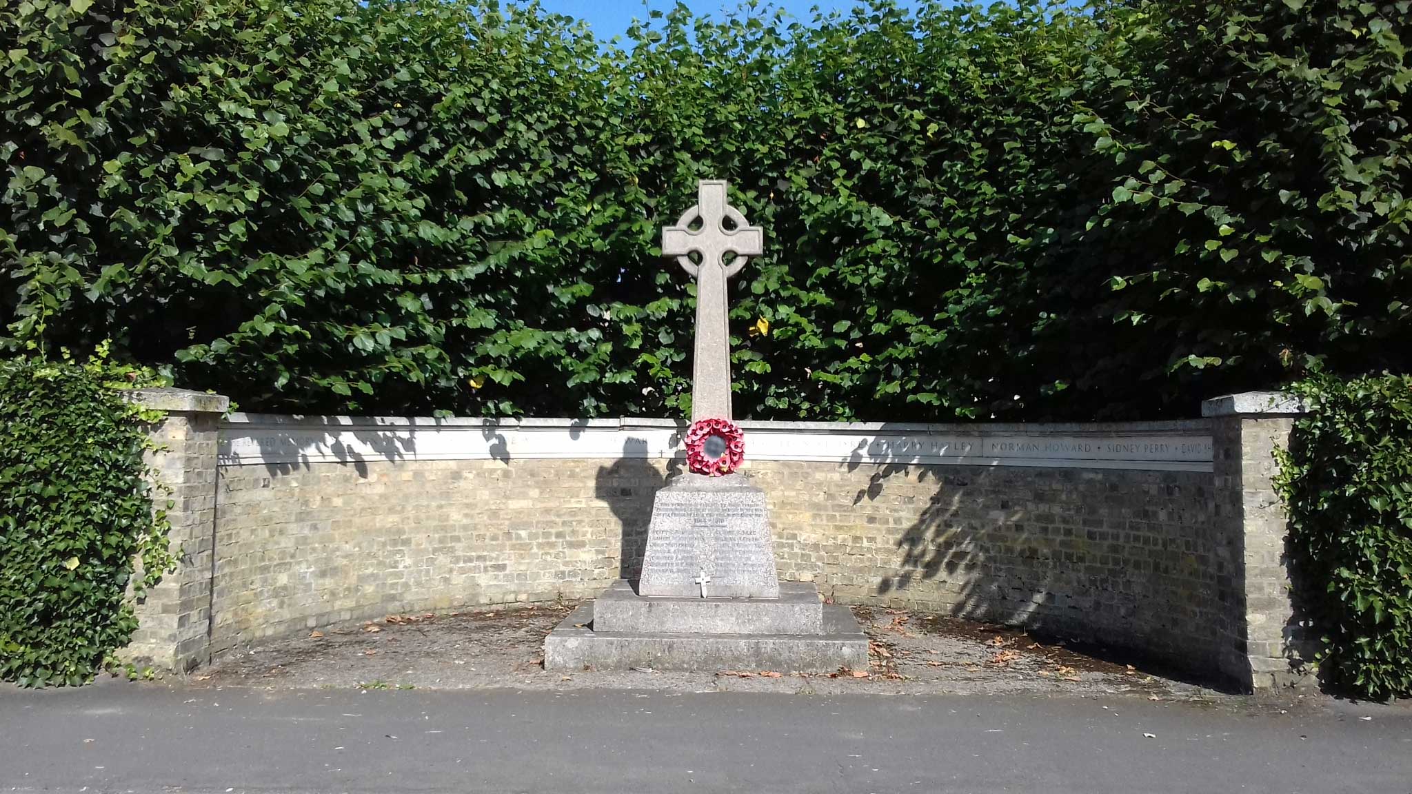 A Celtic cross stands on a tapering plinth in a semi-circular remembrance wall