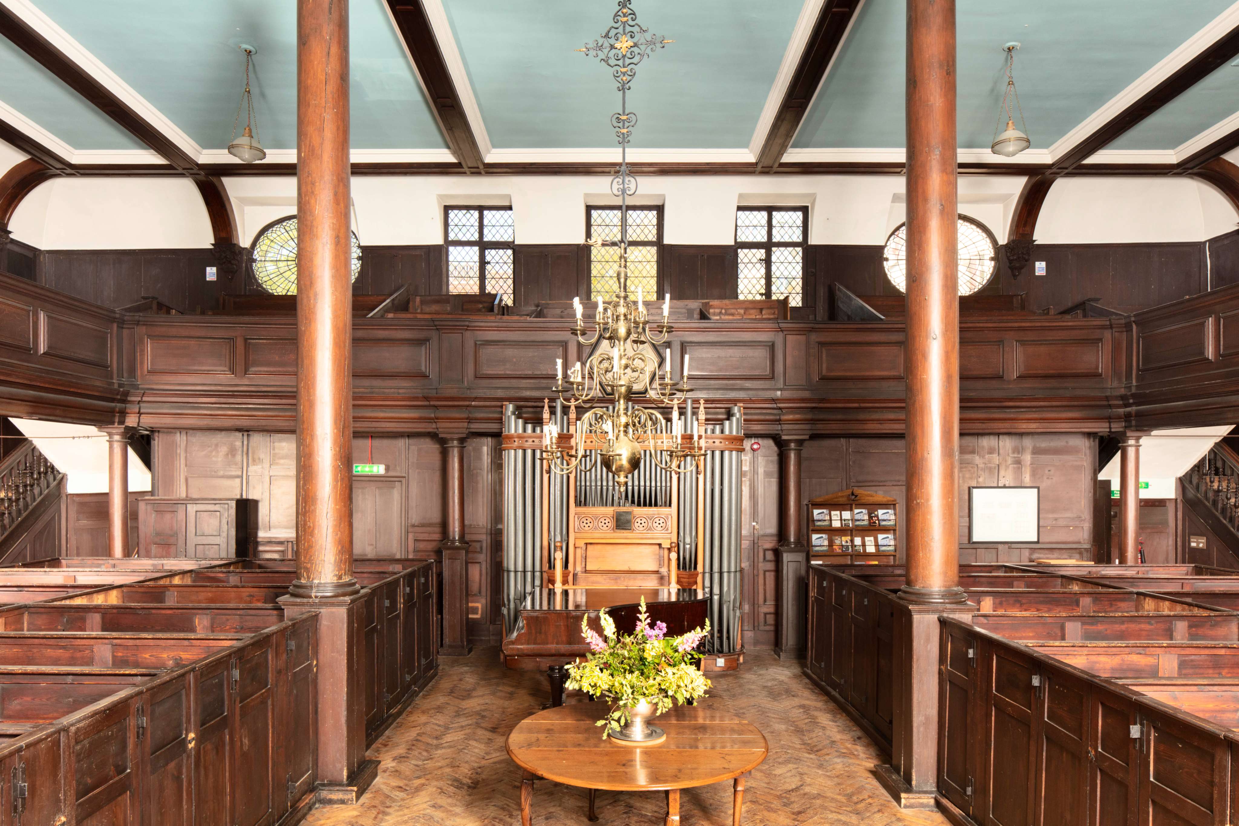 The beautiful interior of the Unitarian Meeting House showing dark wood pews, parquet flooring, wooden pillars and the late 19th century organ. A chandelier hangs from the middle of the ceiling.