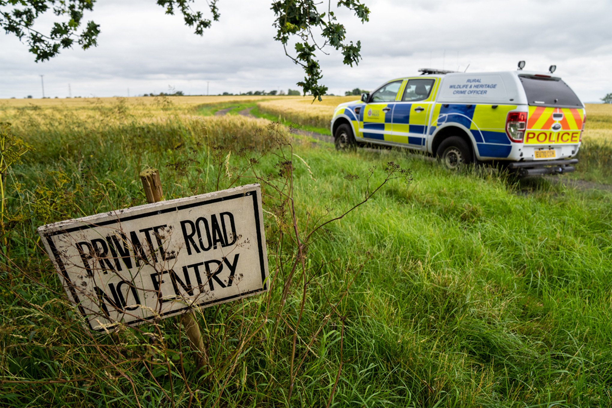 A photograph of a police SUV parked in a field. A sign in the foreground reads "Private Road / No entry"