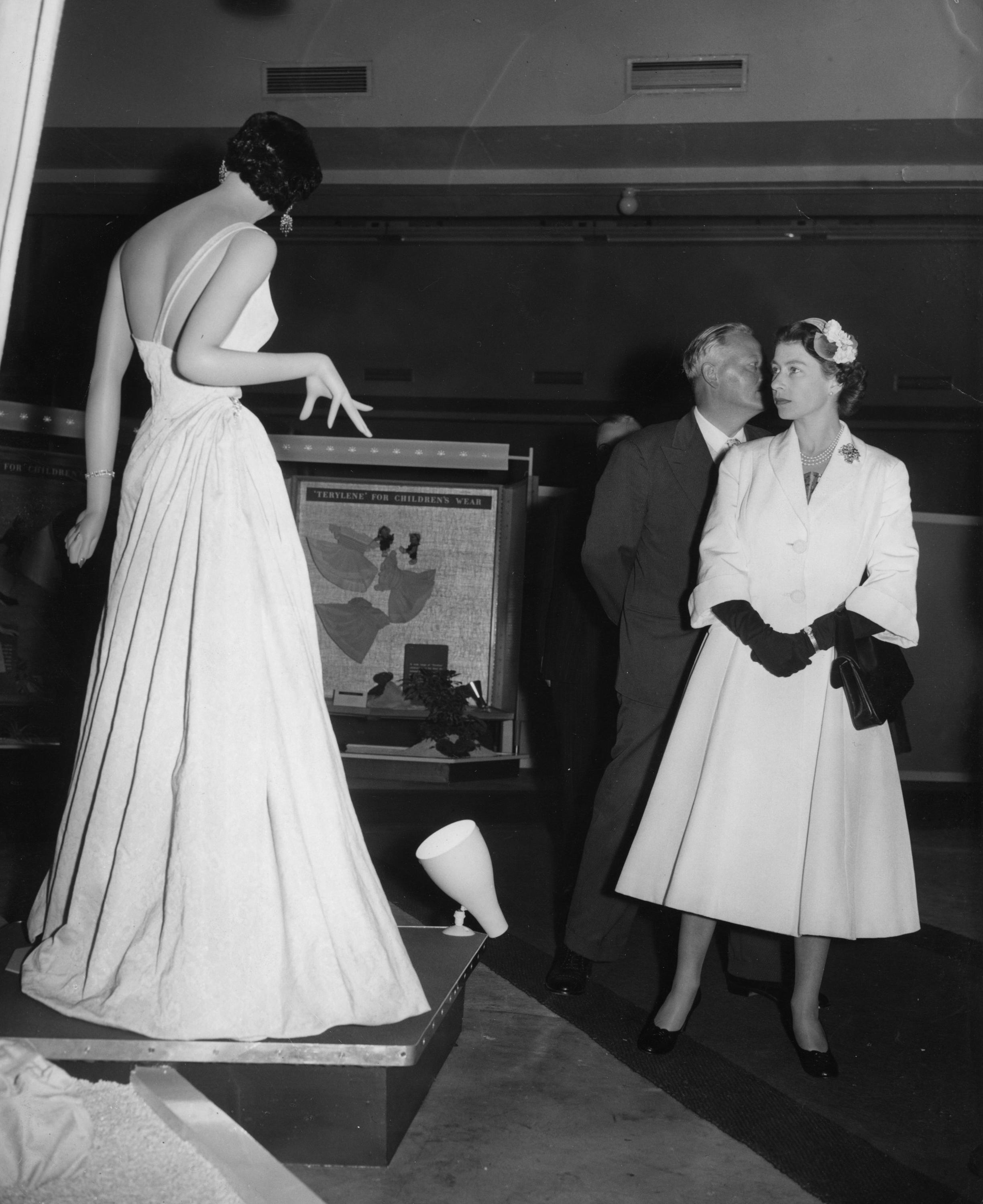 Queen Elizabeth II pauses to look at a dress, made of 100 percent Terylene, as she tours an exhibition of goods made from the synthetic fibre at the Imperial Chemical Industries' works at Wilton, near Middlesbrough, 5 June 1956. © PA Images / Alamy Stock Photo.