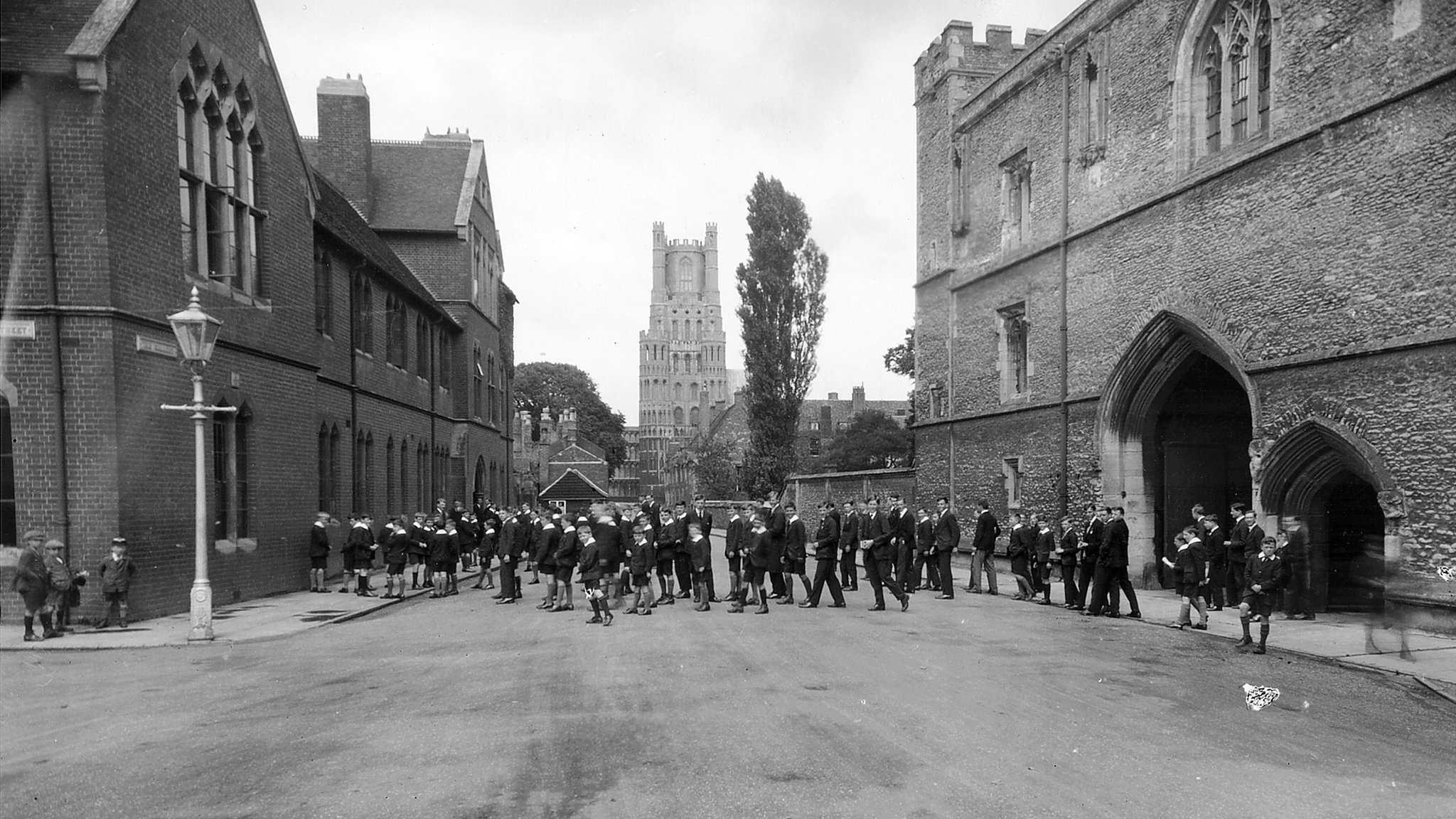 Archive black and white photograph of a group of schoolboys in the street between a brick building and a flint and stone medieval gatehouse, with a church tower in the background.
