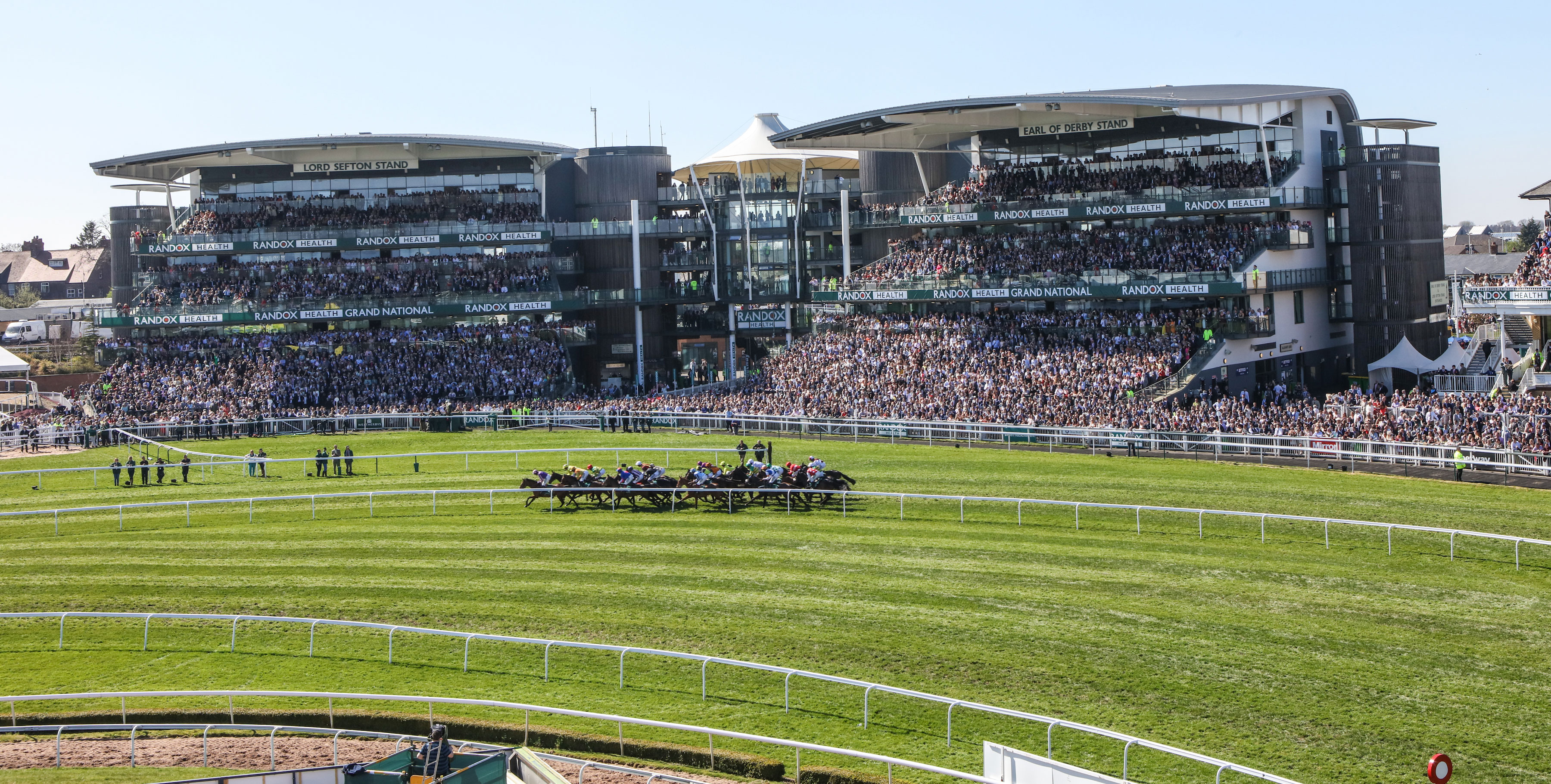 The packed stands on race day at Aintree with a tightly bunched group of horses galloping down the track in the foreground.