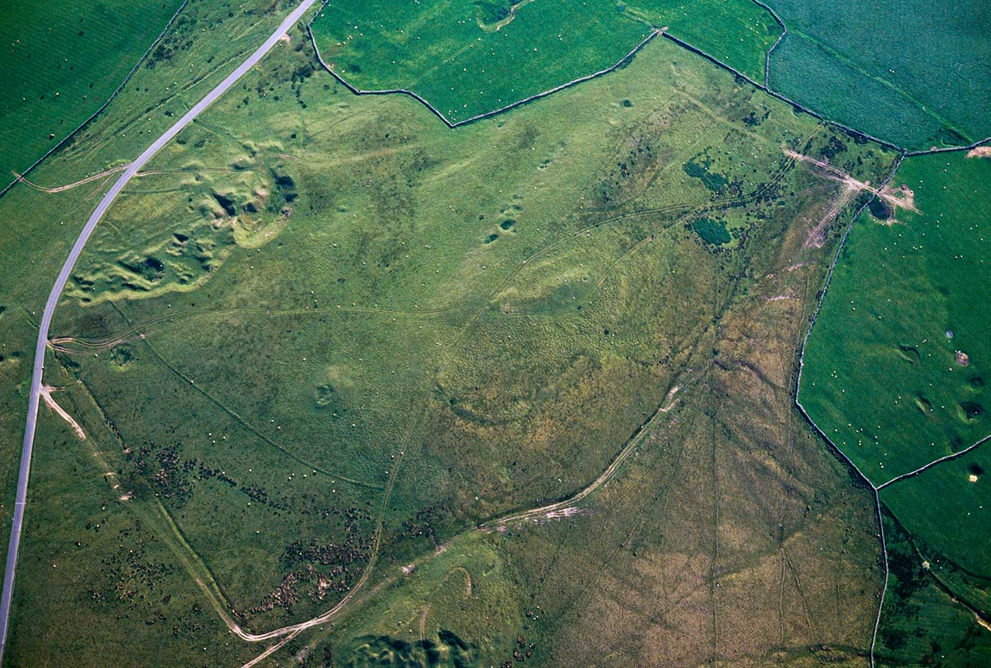 Colour aerial photo showing moorland crossed by tracks, with an oval enclosure on a hilltop and some quarrying to the left