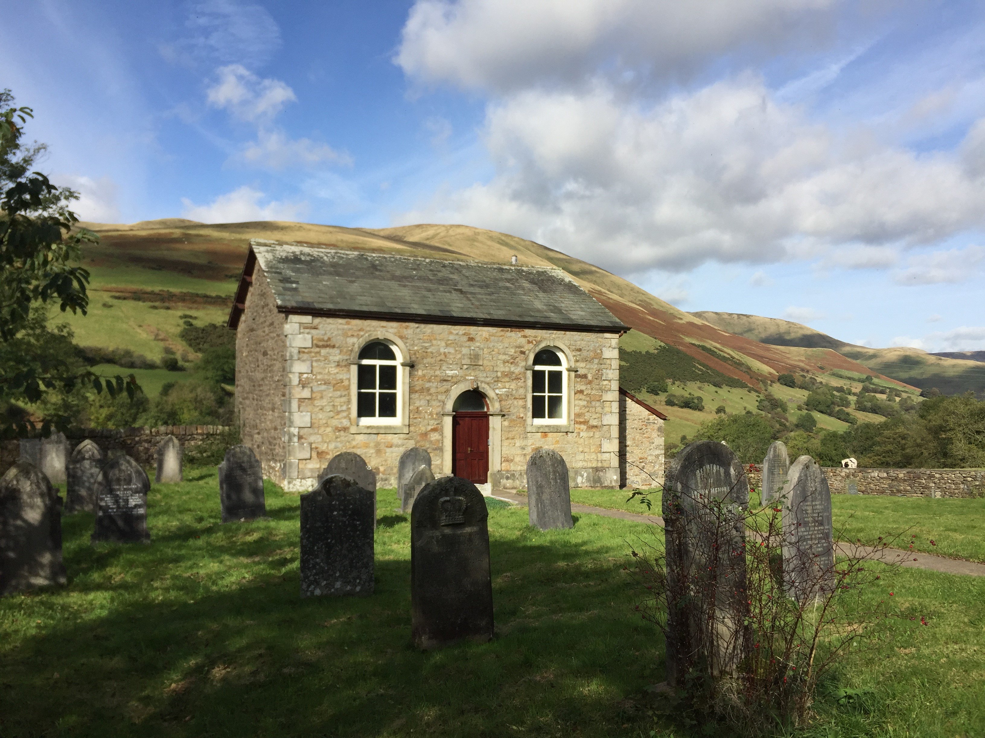 Small stone chapel set in Cumbrian mountain scenery on a sunny day.