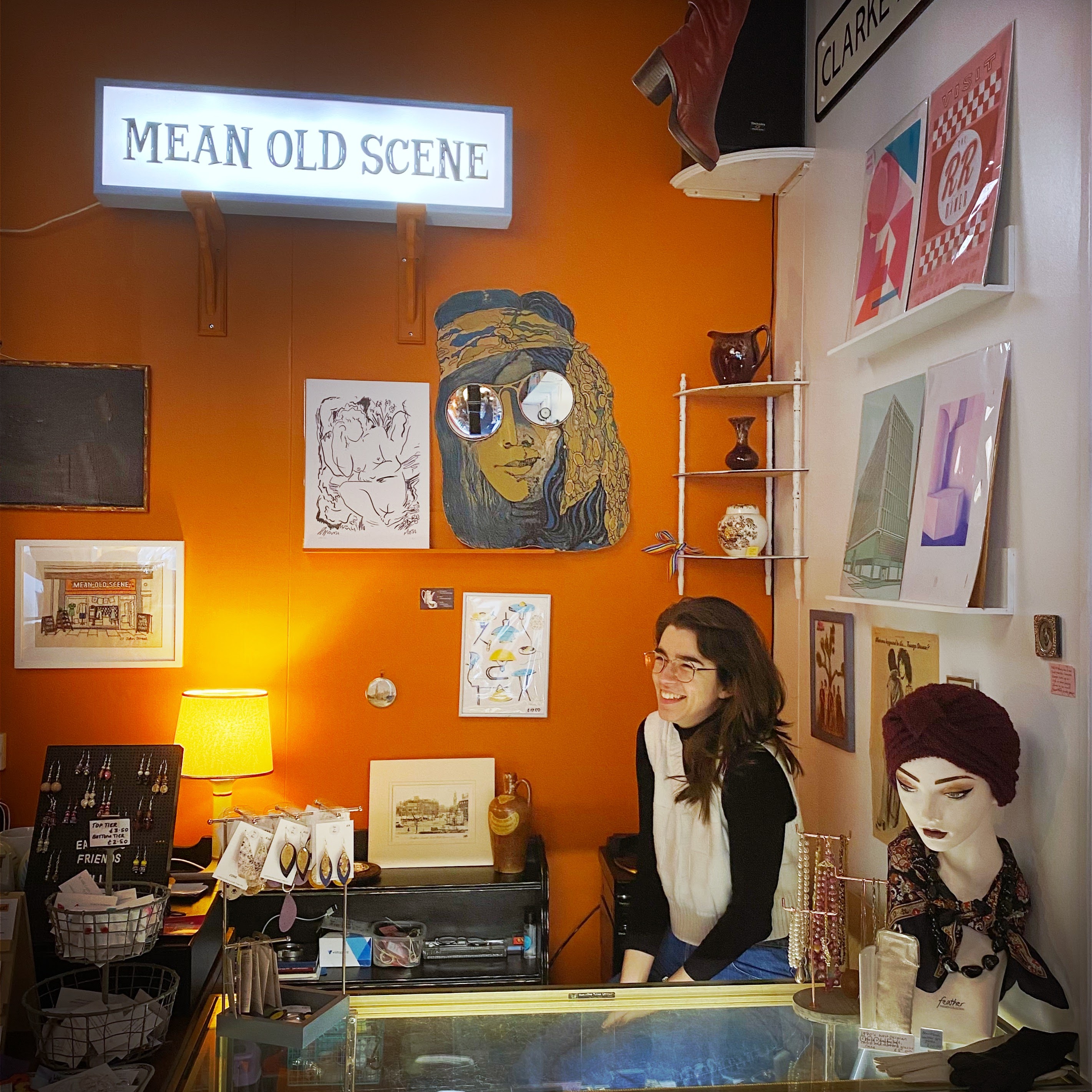 Photograph of a woman with dark hair smiling behind the counter of her store. Jewellery and artwork is on sale around her.