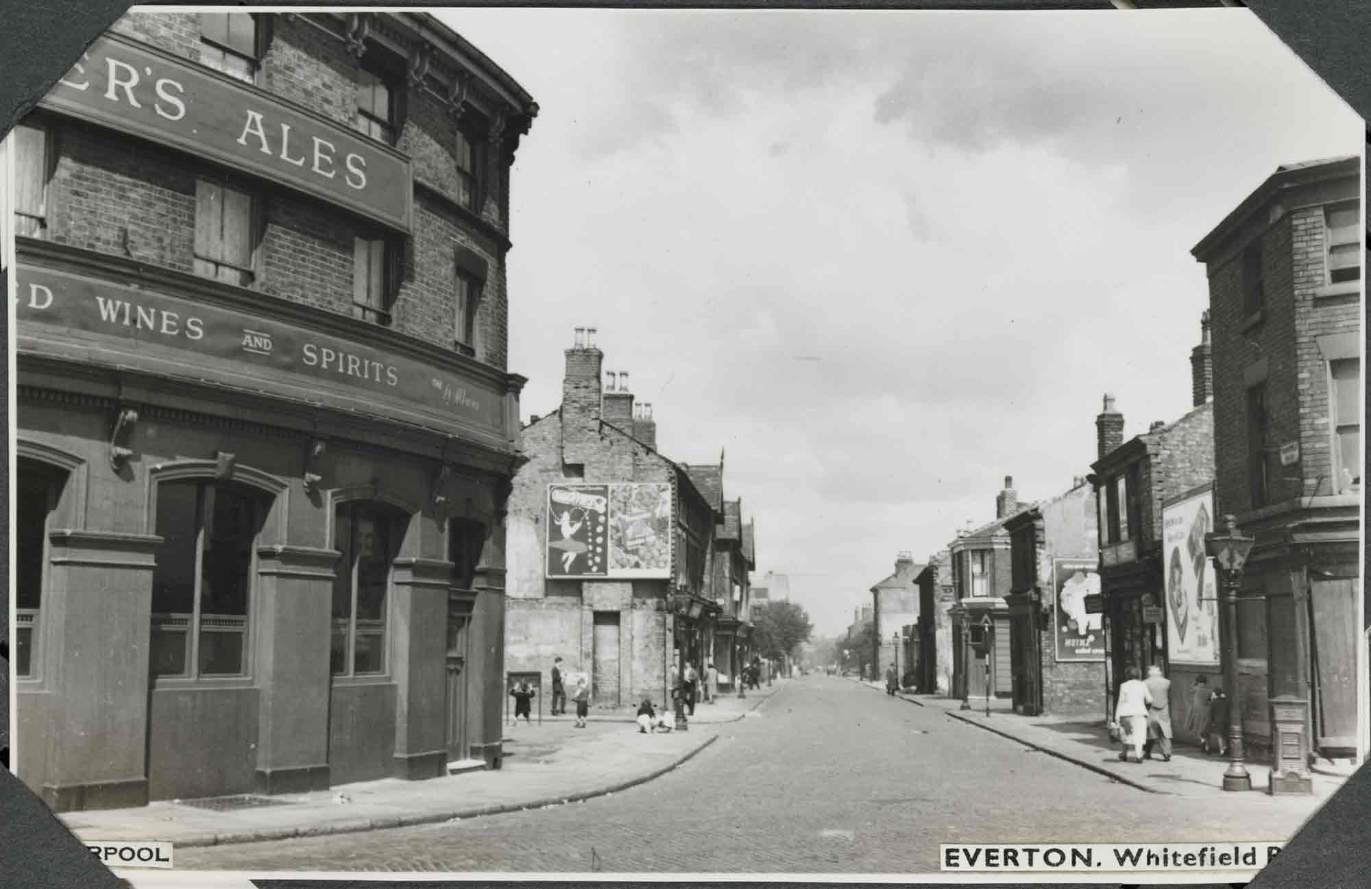 Black and white photograph of an urban street scene that looks down a long, straight road flanked by two- and three-storey brick houses and commercial buildings. In the left foreground, the street and pavement curve in front of a curved, three-storey public house. Beyond is an open patch of land with children playing on it. The exposed end of the next building carries an advertisement board. The buildings on the opposite side are interrupted by empty plots, alleys, or side roads. A label applied to the bottom-right of the photograph reads: 'Everton. Whitefield Road' and a label applied to the bottom-left reads: 'Liverpool', parts of which are obscured by corner photograph album mounts.