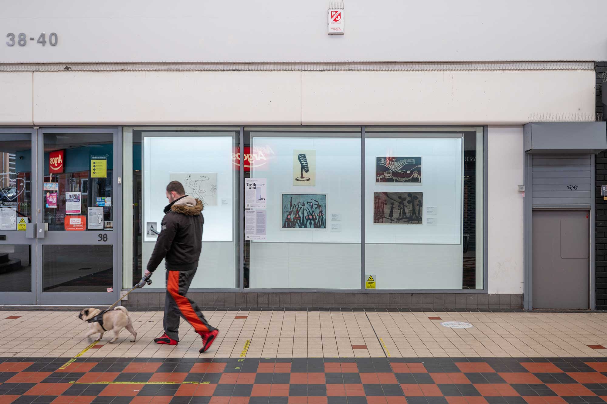 Man walking a dog past a shop where prints and drawing are displayed in the window