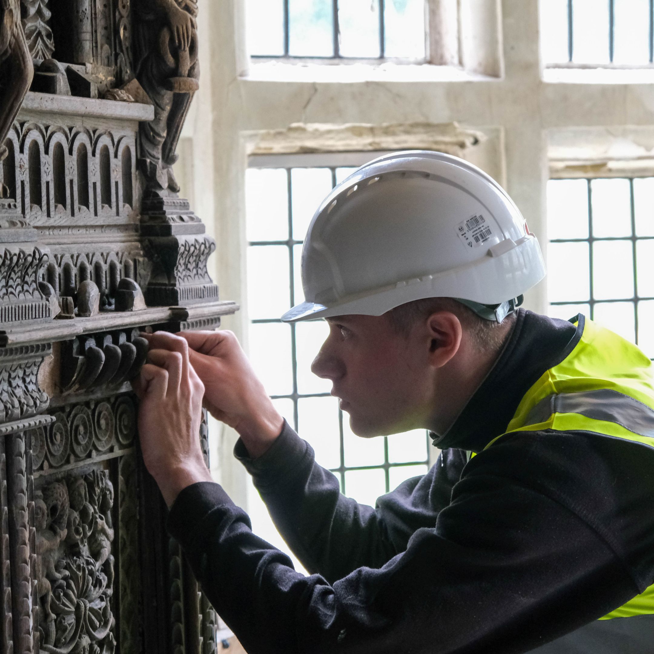 A photograph of a man in a white helmet and hi-vis jacket closely inspecting a decorative wooden object in front of a window.
