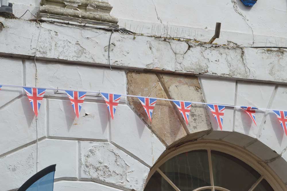 A photograph showing a detail of an external wall with Union Jack bunting across it. The white-rendered stonework over a round arched window shows signs of decay and poor repair.