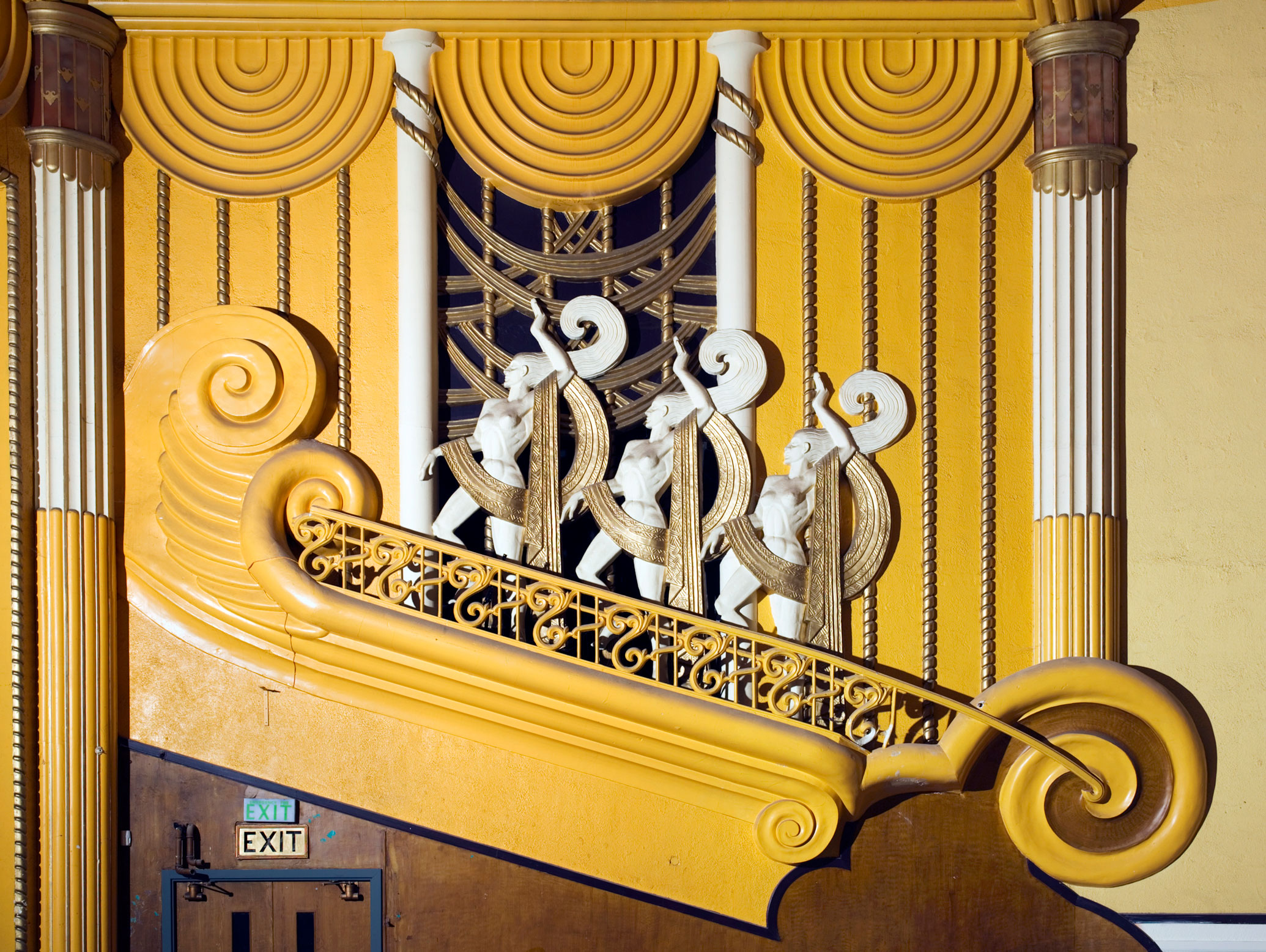 A detail of plaster decoration in vibrant yellow and white, including a row of three mythical figures in an art deco style.