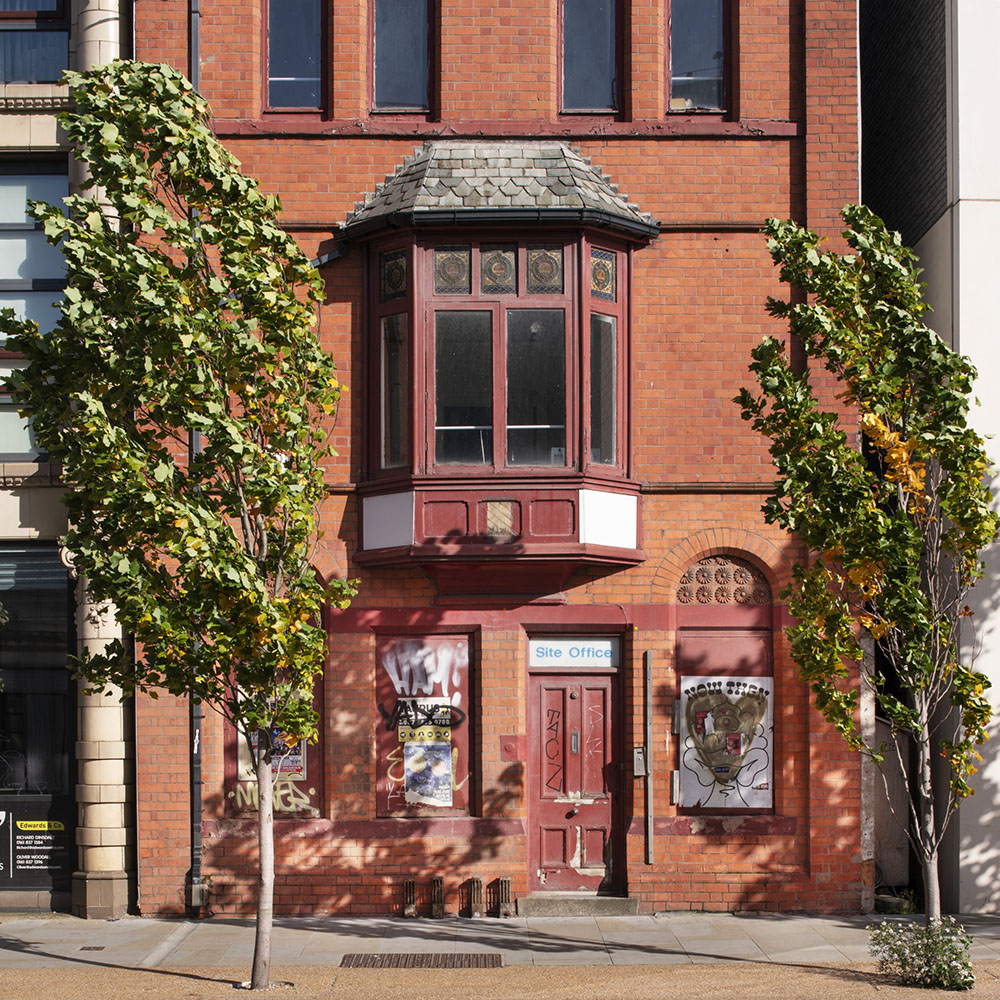 A red brick three-storey townhouse, with saplings in front, and red wooden detailing.