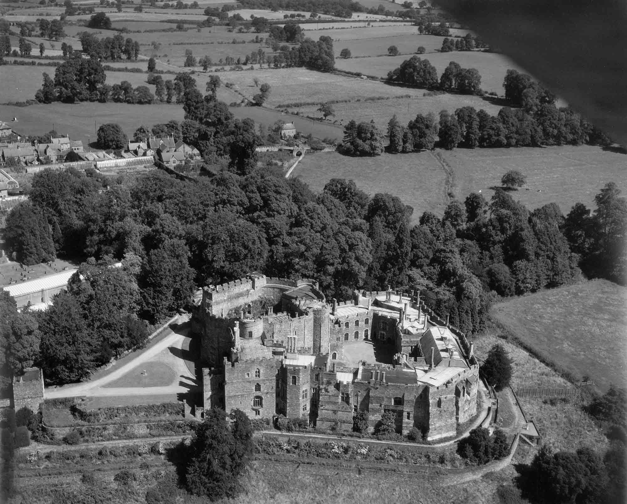 Black and white oblique aerial photograph featuring in the foreground a stone castle with crenelated walls and inner and outer courtyards. Tall trees extend across the centre of the photograph. Adjacent to the castle and stretching beyond the trees are fields bounded by hedges and clusters of trees. In the left middle distance are houses, gardens and a narrow bridge.