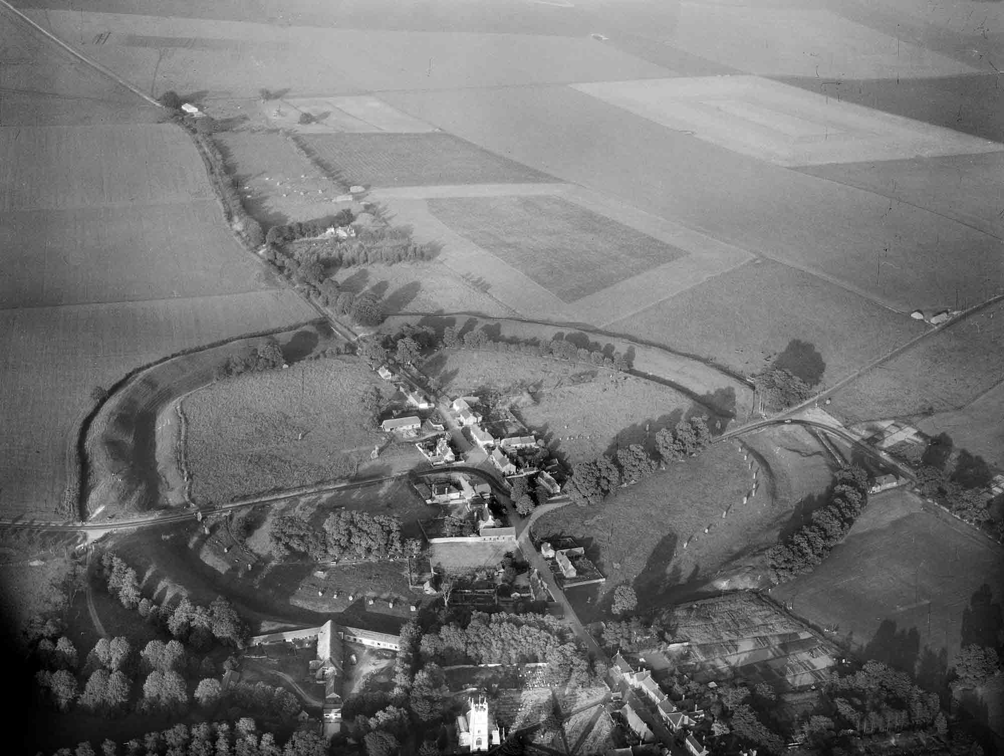 Black and white oblique aerial photograph of part of a village and a circular henge enclosure featuring inner and outer ditches on either side of a grassy bank. The upper half of the photograph features multiple fields and a tree-lined road or track heading into the distance.