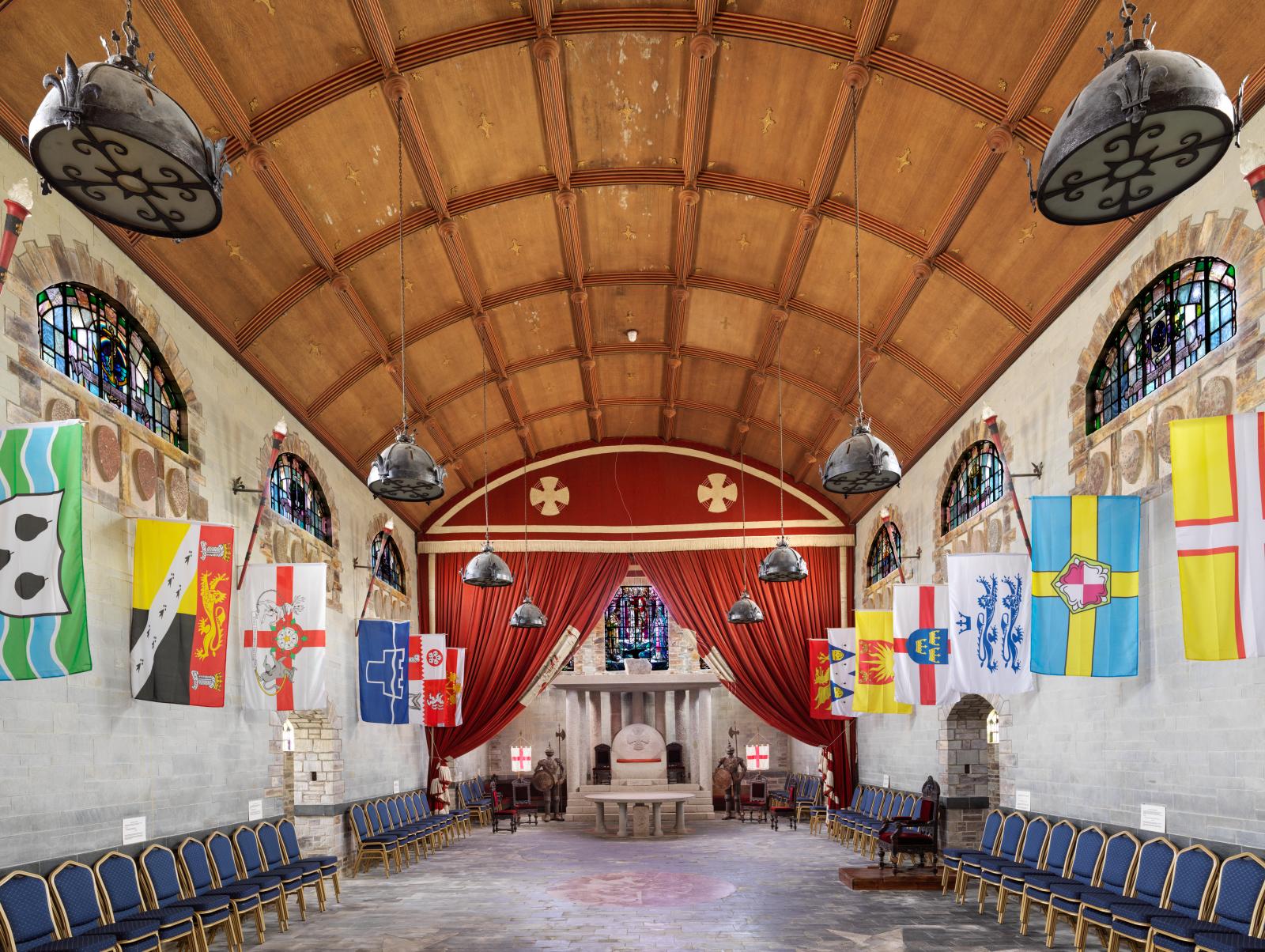 A large hall space with stone walls and a wooden barrel-vaulted ceiling. Stained glass windows and hung banners and curtains add colour to the space.