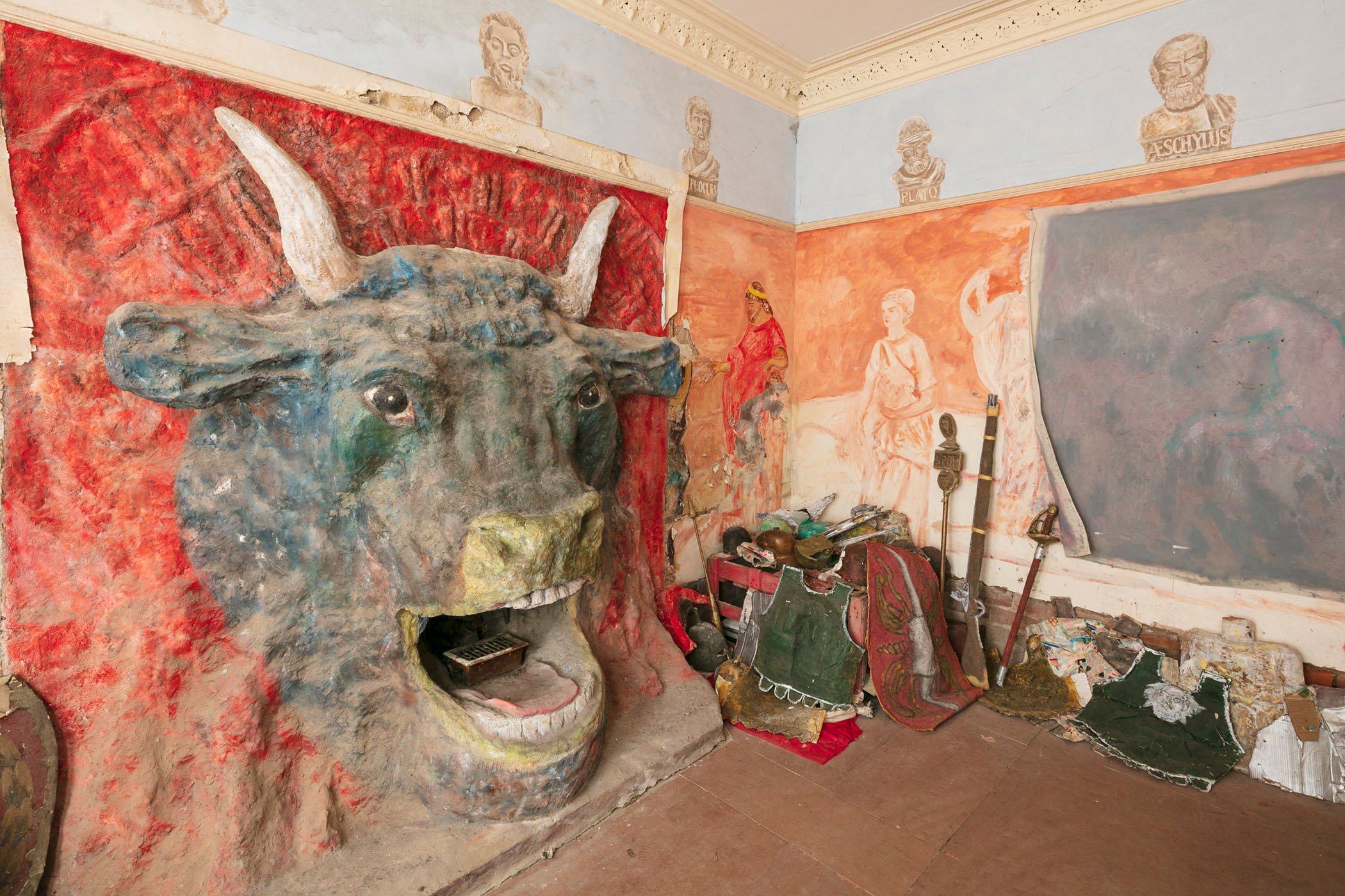 A photograph of a room containing a large Minotaur head sculpture and Roman-Greco-inspired artwork painted on the walls.