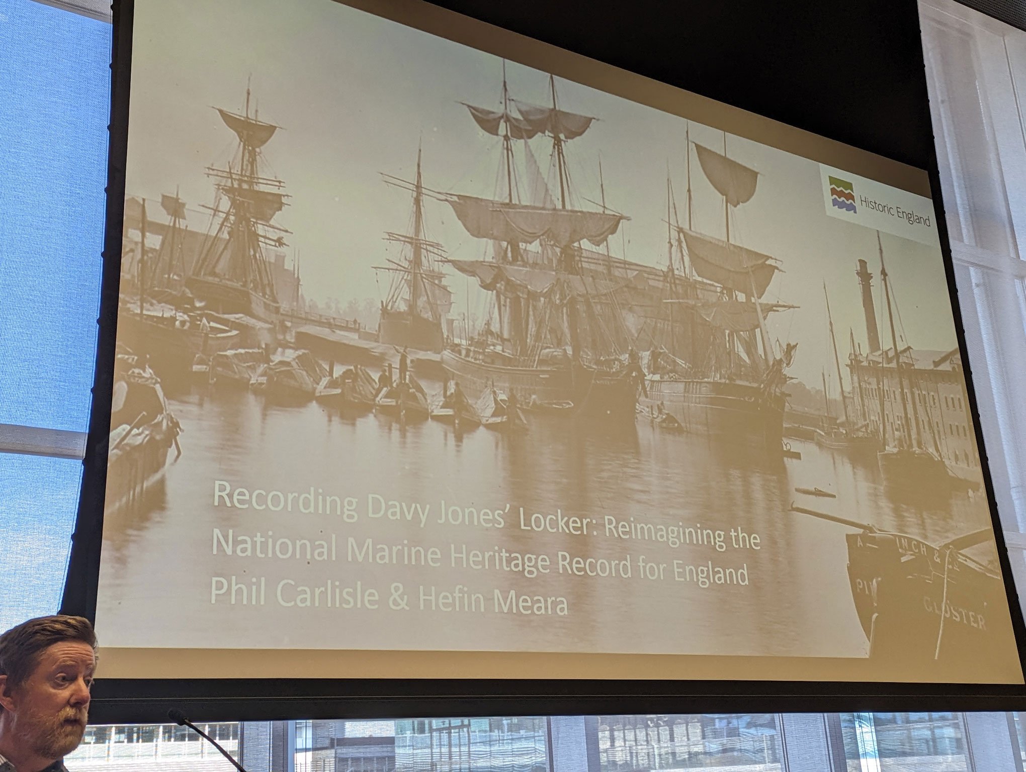 Photograph of a man presenting at a conference, he is standing in front of a screen upon which is projected an archive image of a harbour with sailing vessels.