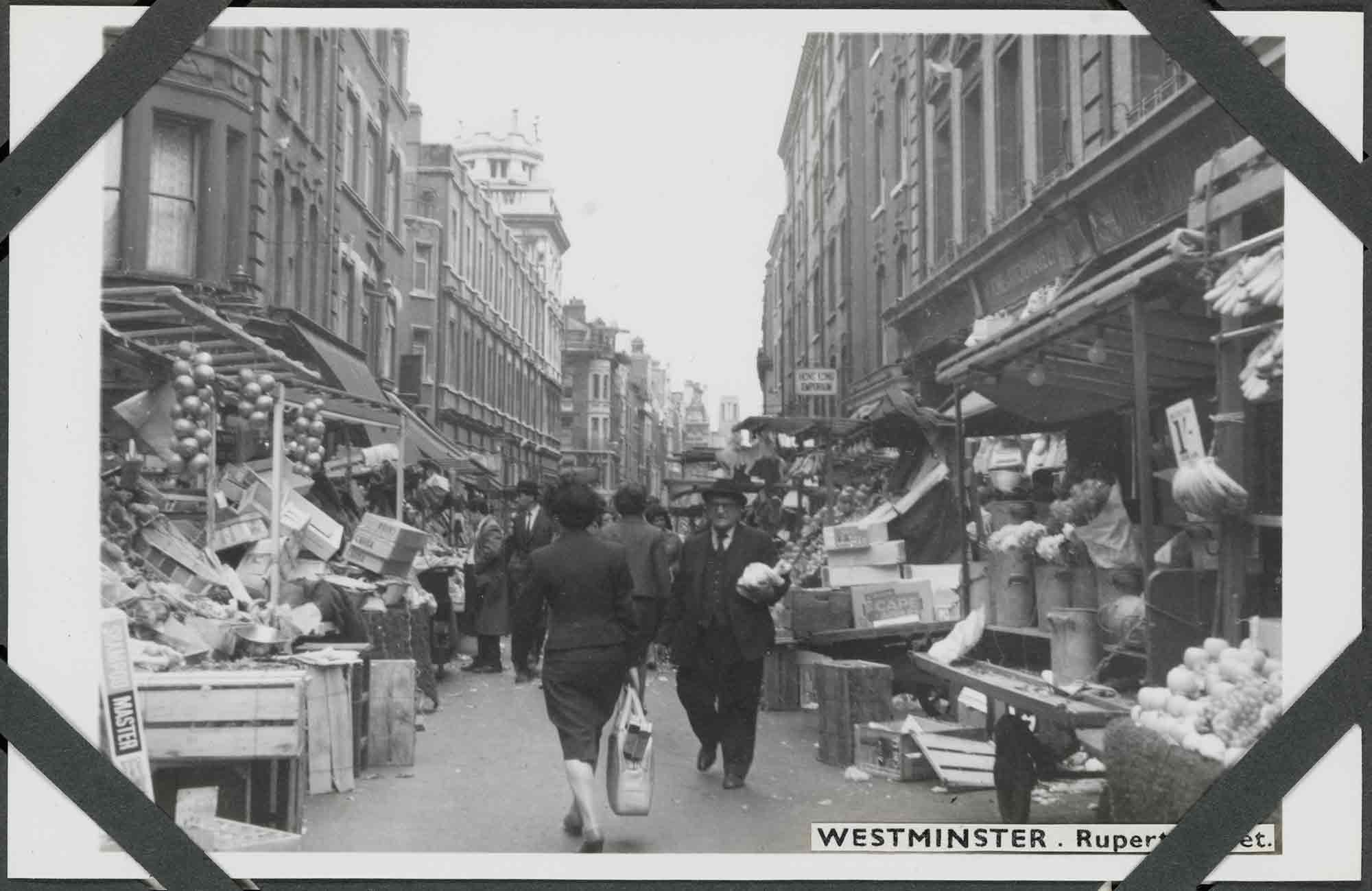 Black and white photograph of an urban street scene featuring people walking on a road flanked by market stalls set out in front of rows of stone-fronted buildings of multiple storeys. The market stalls include displays of fruit, vegetables, and flowers. Several wooden boxes are discarded around them. In the foreground, a man in a dark three-piece suit and hat walks toward the camera carrying a bag, a woman in a dark two-piece suit and carrying a light shopping bag walks away from the camera. Four corner photograph album mounts are visible. 