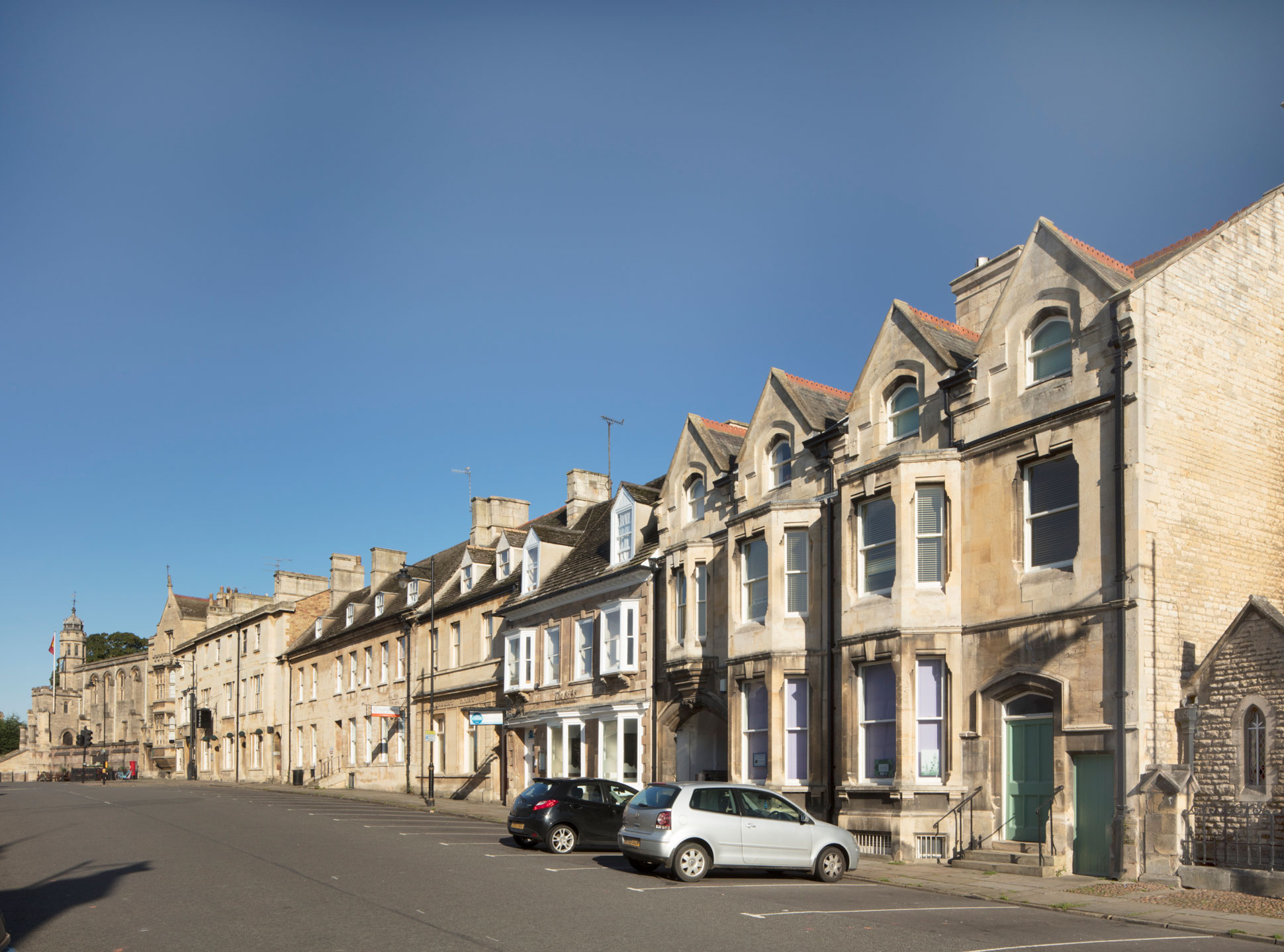 Colour photo showing a view along a row of three-storey 19th century buildings built in a light ashlar stone. Parking bays line the road in front of the buildings. Two cars are parked out front.