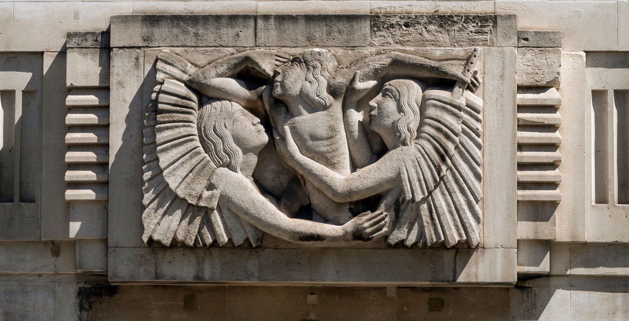 A relief sculpture on a building depicting two winged figures embracing a central figure who  is carrying a flute.