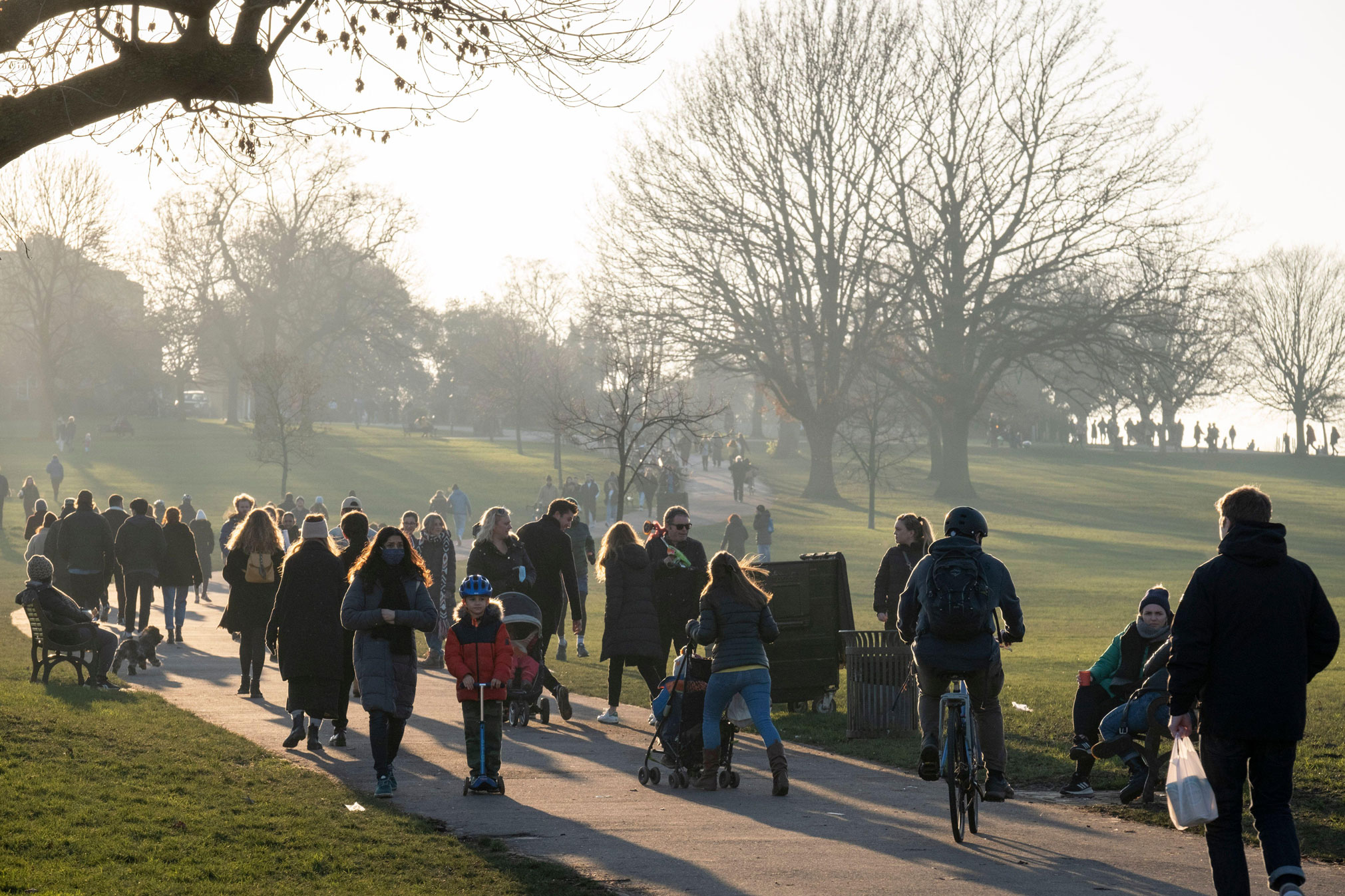 Lots of people, including children, walking, scootering, cycling and running along a path through grass and trees in a park. 