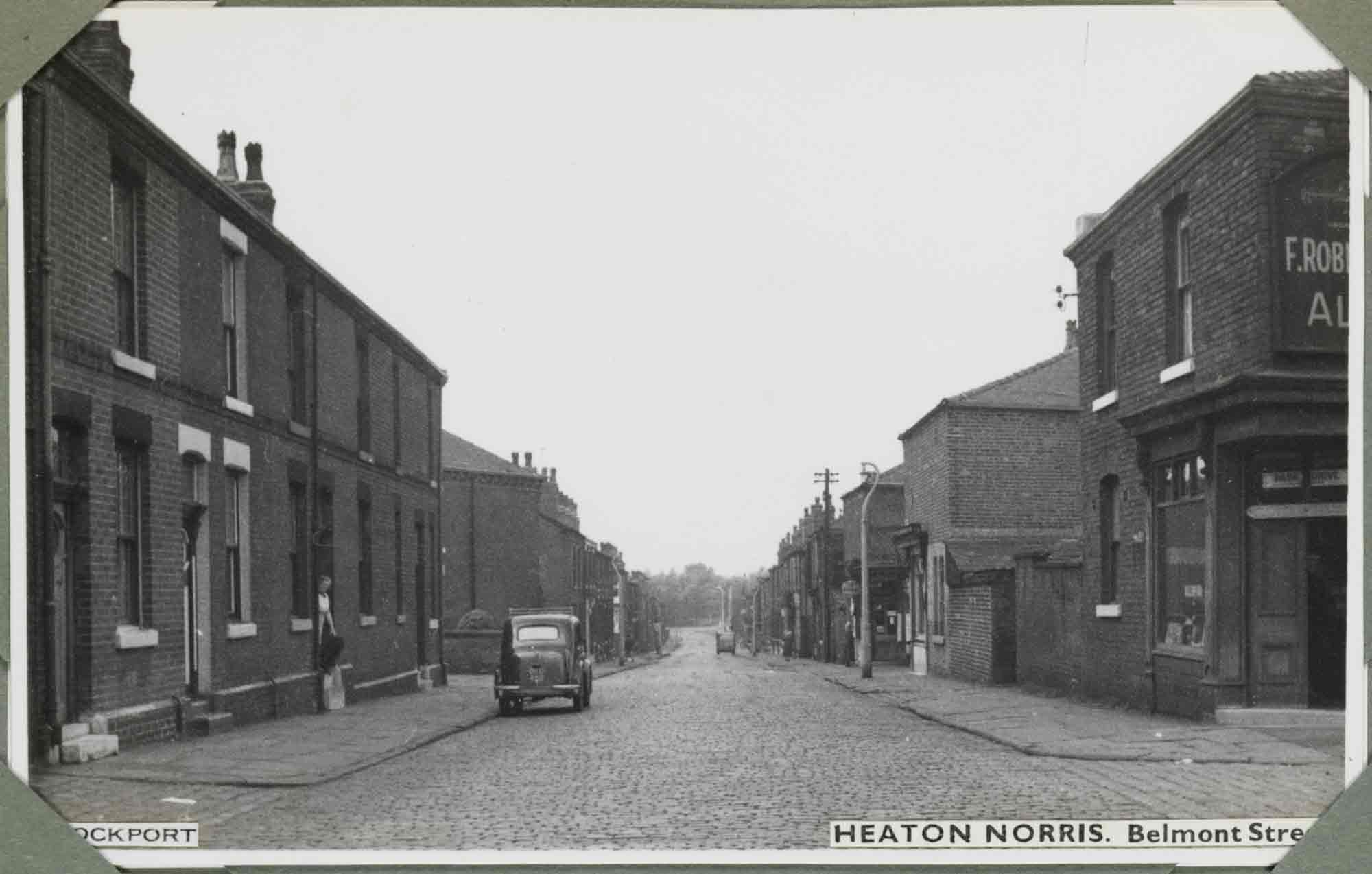 Black and white photograph of an urban street scene. The view looks straight along a cobbled road, gently sloping into the distance. The street is empty apart from a couple of parked cars and a woman on the doorstep of a brick terrace house to the left side of the street. The two-storey buildings flanking the street are mostly houses that front directly onto the pavement. On the right side, some commercial premises can be made out, the one in the foreground having a double-door corner entrance. A label applied to the bottom-right reads: 'Heaton Norris. Belmont Street' and a label applied to the bottom-left reads: 'Stockport', although they are partly obscured by corner photograph album mounts. 