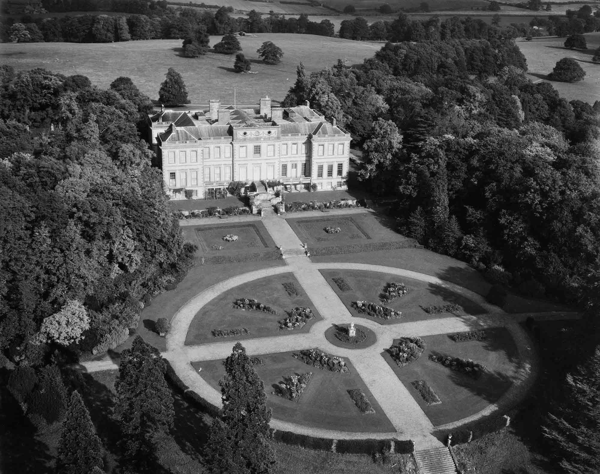 Black and white oblique aerial photograph taken at low altitude. The view shows the garden front of a large country house with parkland and trees beyond. The bulk of the photograph shows a formal garden arrangement, comprising two terraces, the second with sunken lawns divided by a gravel path that extends towards a rose garden enclosed by a circular walk and divided into four segments by cruciform gravel walks. Dense clusters of trees flank the house and surround the gardens.