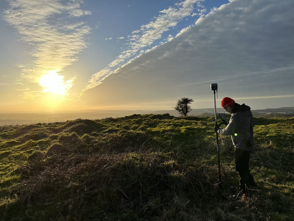 An atmospheric photograph showing a man holding a surveying reflector staff at a historic upland site, lit by low sunlight.