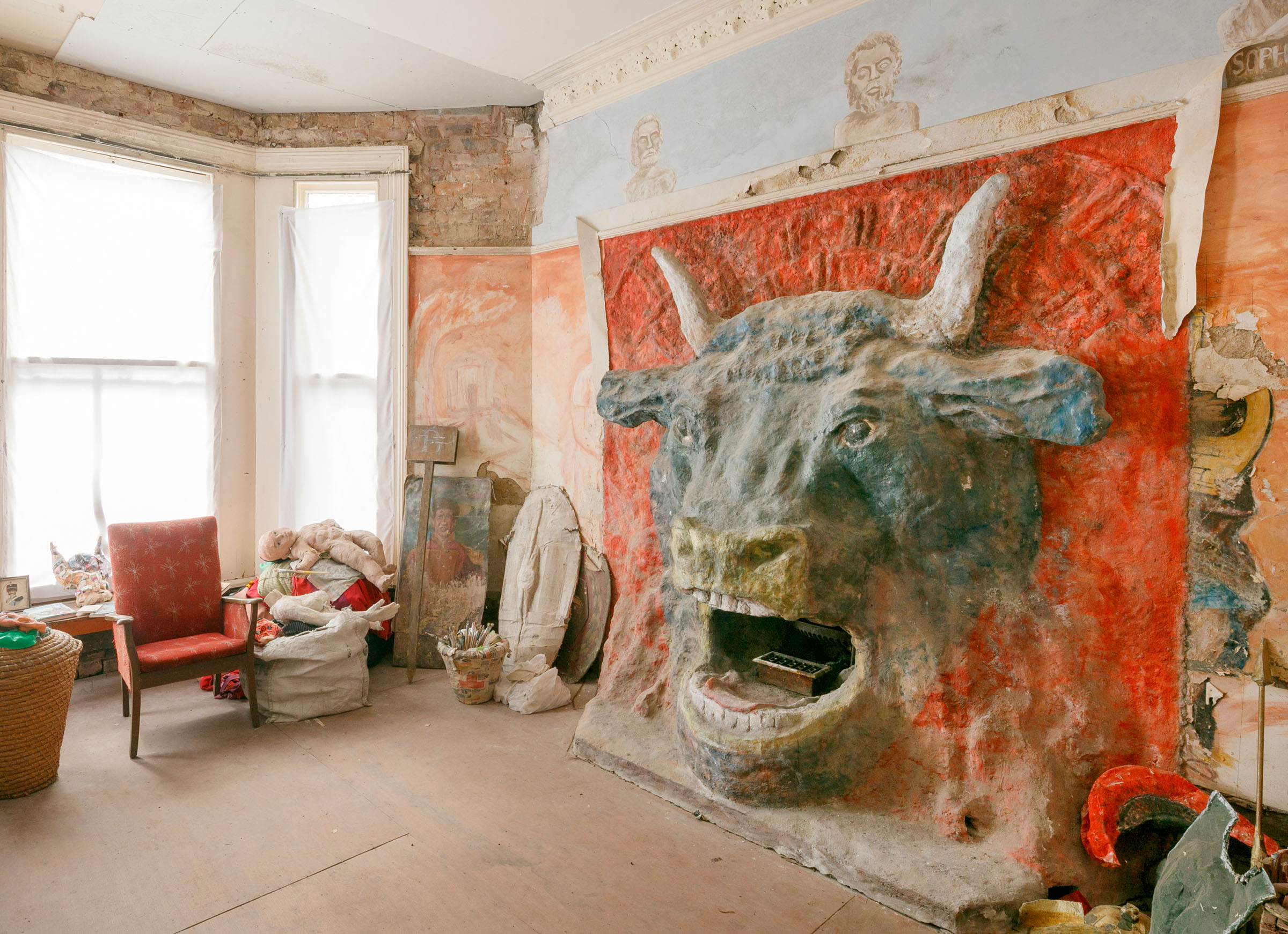 A photograph of a room containing a large Minotaur head sculpture, a chair, a bay window and Roman-Greco-inspired artwork painted on the walls.